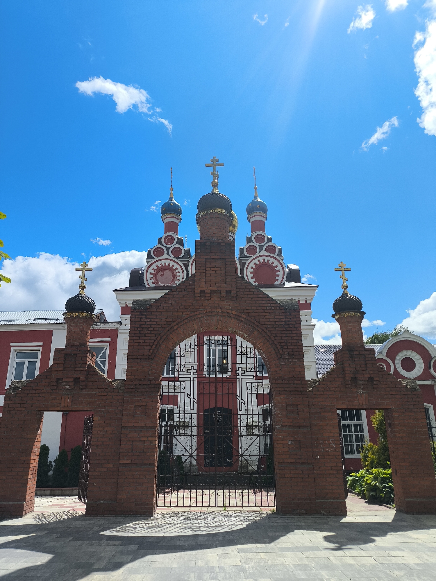 The image captures the entrance of the Church of the Resurrection of Christ in Taldom, Russia. A red brick archway frames the view of the church's facade. The church itself is painted in white and red, with distinctive onion domes topped with golden crosses. The sky above is a vibrant blue, dotted with white clouds and illuminated by bright sunlight. The foreground shows a paved walkway with intricate patterns, and the shadows cast by the archway and foliage suggest it is a sunny day. The overall scene conveys a sense of architectural grandeur and peaceful religious atmosphere.
