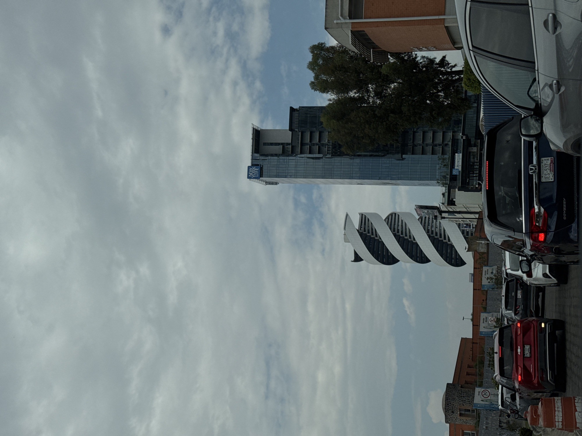 The image shows a street scene in San Bernardino Tlaxcalancingo, Mexico. A line of cars, including a red Chevrolet, a dark blue Honda Odyssey, and a light gray minivan, are stopped in traffic. A modern, spiraling white building stands to the left of the traffic. In the background are a tree, a tall glass building, and other buildings. The sky is mostly cloudy.