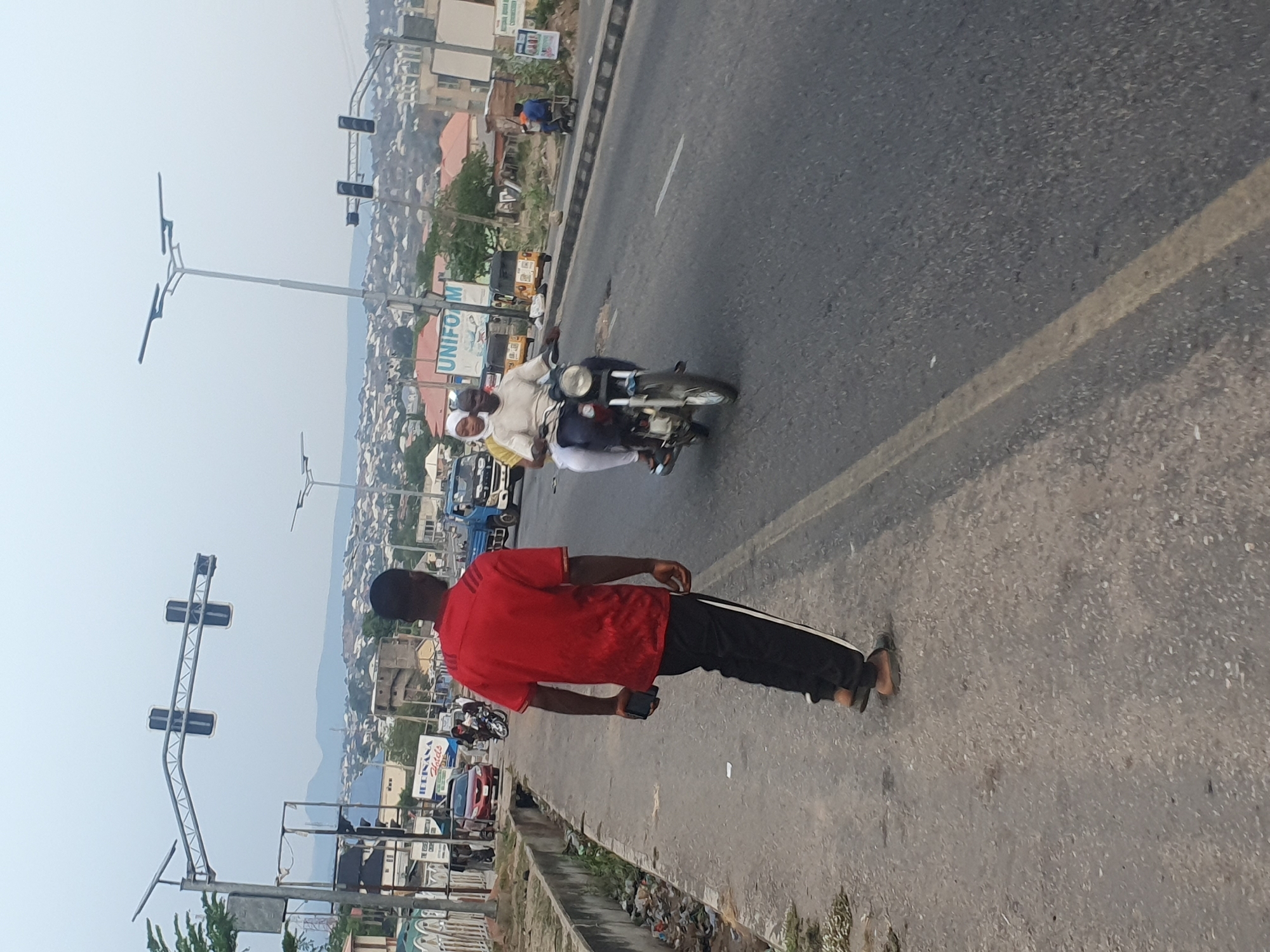 A daytime roadside scene in Lokoja, Nigeria, features a man in a red shirt and dark pants walking on the sidewalk. He is holding a small, dark object in his hand. To his left is a busy road with vehicles, including a motorcycle carrying two people. Buildings and trees are visible in the background, along with traffic lights and streetlights. The sidewalk is littered with debris.