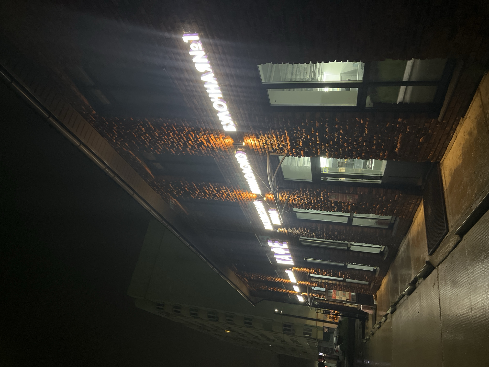The image depicts an urban street scene at night in the city of Chaikovsky, Russia, following or during light rain. The ground, including the sidewalk and parts of the street, is wet with visible reflections.
In the foreground, a two-story commercial building with a dark reddish-brown, textured brick facade runs diagonally across the frame from left to right. The building features multiple large, rectangular windows with dark frames, and several entrance doors. Mounted on the facade are white, illuminated signs in Cyrillic script. From left to right, these signs read: "КАФЕ" (CAFE), "МАЙОРСКАЯ КУХНЯ" (MAYORSKAYA KUKHNYA), and "БУЛОЧНАЯ №1" (BAKERY №1). Several warm, orange-yellow spotlights are mounted on the facade, illuminating the brickwork between the windows and below the signs. A light-colored gutter system is visible along the roofline.
The sidewalk in front of the building is wet, showing ripples and reflections of the building's lights. Dark doormats are visible at building entrances. A low curb separates the sidewalk from a narrow strip of ground and the street. In the distant left, a light-colored vehicle is partially visible parked on the street. A tall, multi-story residential apartment building with a light green or pale yellow facade and numerous windows stands in the background on the left. The sky is dark and overcast. There are no people visible in the image.