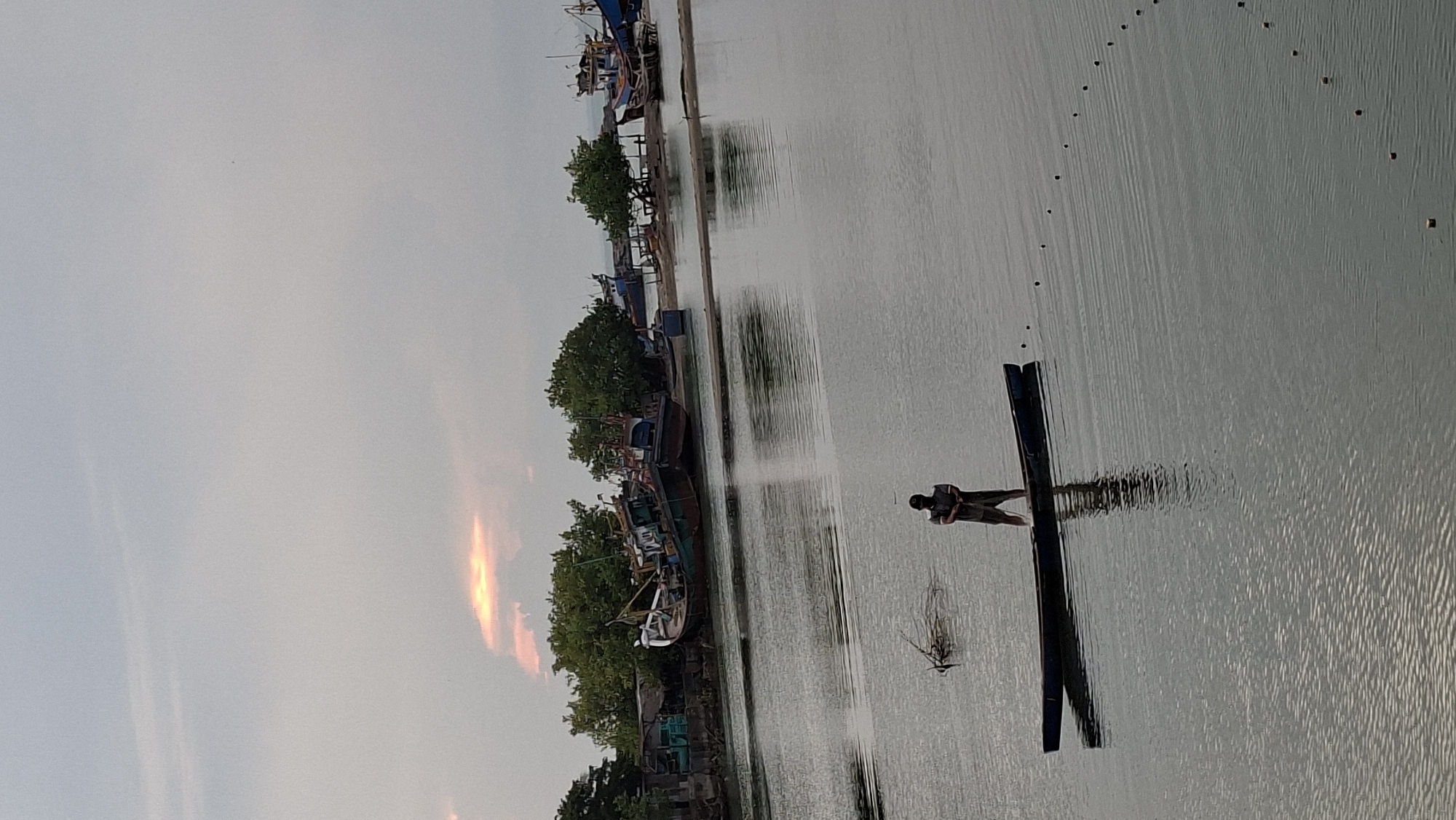 The image depicts a serene waterscape, likely in Lhokseumawe, Indonesia. In the foreground, a lone individual stands in a narrow, dark boat, casting a net or fishing line into the calm water. The water reflects the overcast sky, with subtle ripples disturbing the surface. Further in the background, several fishing boats are moored near a vegetated shoreline. The boats appear to be of varying sizes and conditions, some with visible structures and rigging. The shoreline itself is characterized by lush green trees and some rudimentary structures, suggesting a coastal or riverine settlement. The sky is a soft gray, with a hint of diffused sunlight breaking through the clouds in the upper left portion of the image, casting a warm glow on the clouds. The overall atmosphere is tranquil and suggests a quiet moment in the day, possibly early morning or late afternoon.