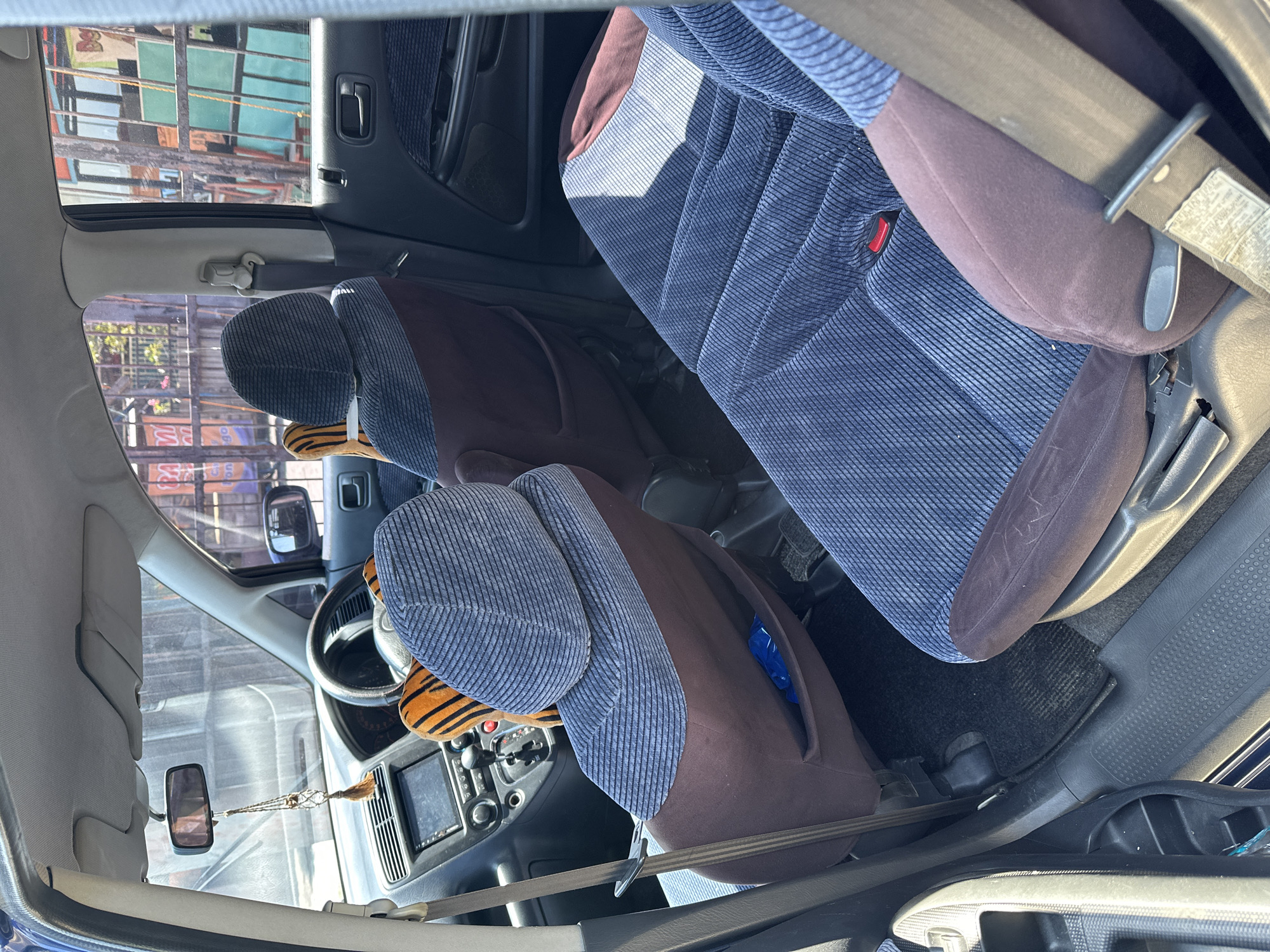 This image, taken in the city of Bulakrejo, Indonesia, shows the interior of a blue car. The focus is on the rear passenger seats, which are upholstered in a corduroy-like fabric with blue stripes and dark brown accents. A blue plastic bag is visible in a pocket on the back of the driver's side rear seat. In the front of the car, a dashboard with a navigation screen and gear shift is visible. Two tiger-striped plush toys are attached to the headrests of the front seats. The windows of the car offer a view of an outdoor area with a fence and some buildings, suggesting an urban or semi-urban setting. The lighting indicates it is daytime. There are no people visible in the car.