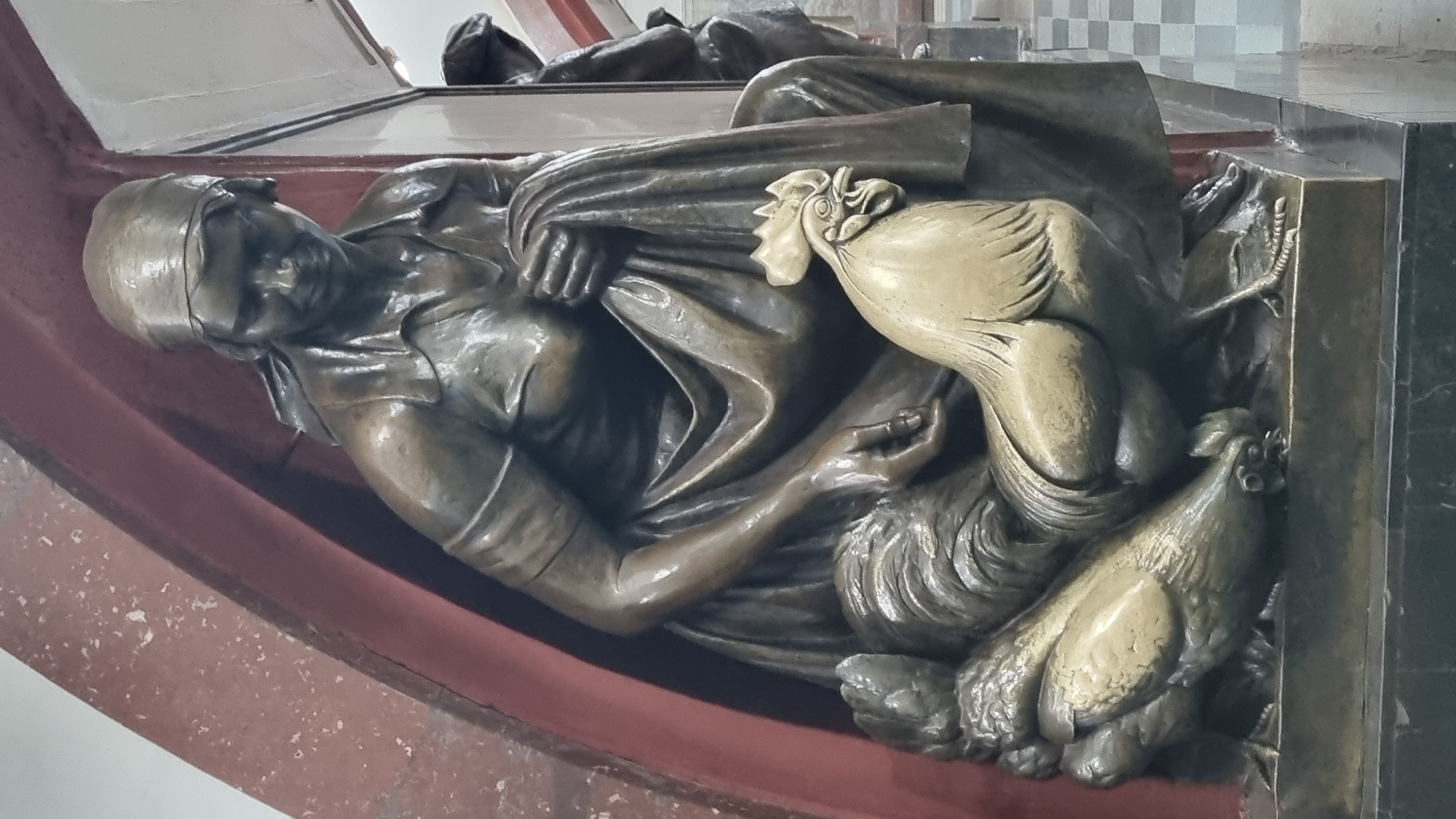 The image shows a bronze sculpture of a woman in a headscarf and apron, holding a piece of cloth. She is seated, with a rooster in the foreground and chickens beside her. The sculpture is set against a red archway. In the background, there is a checkered floor and another bronze sculpture of a person. The sculpture appears to be located indoors, possibly in a public building or transit station. The overall lighting is dim, with highlights on the bronze surfaces.