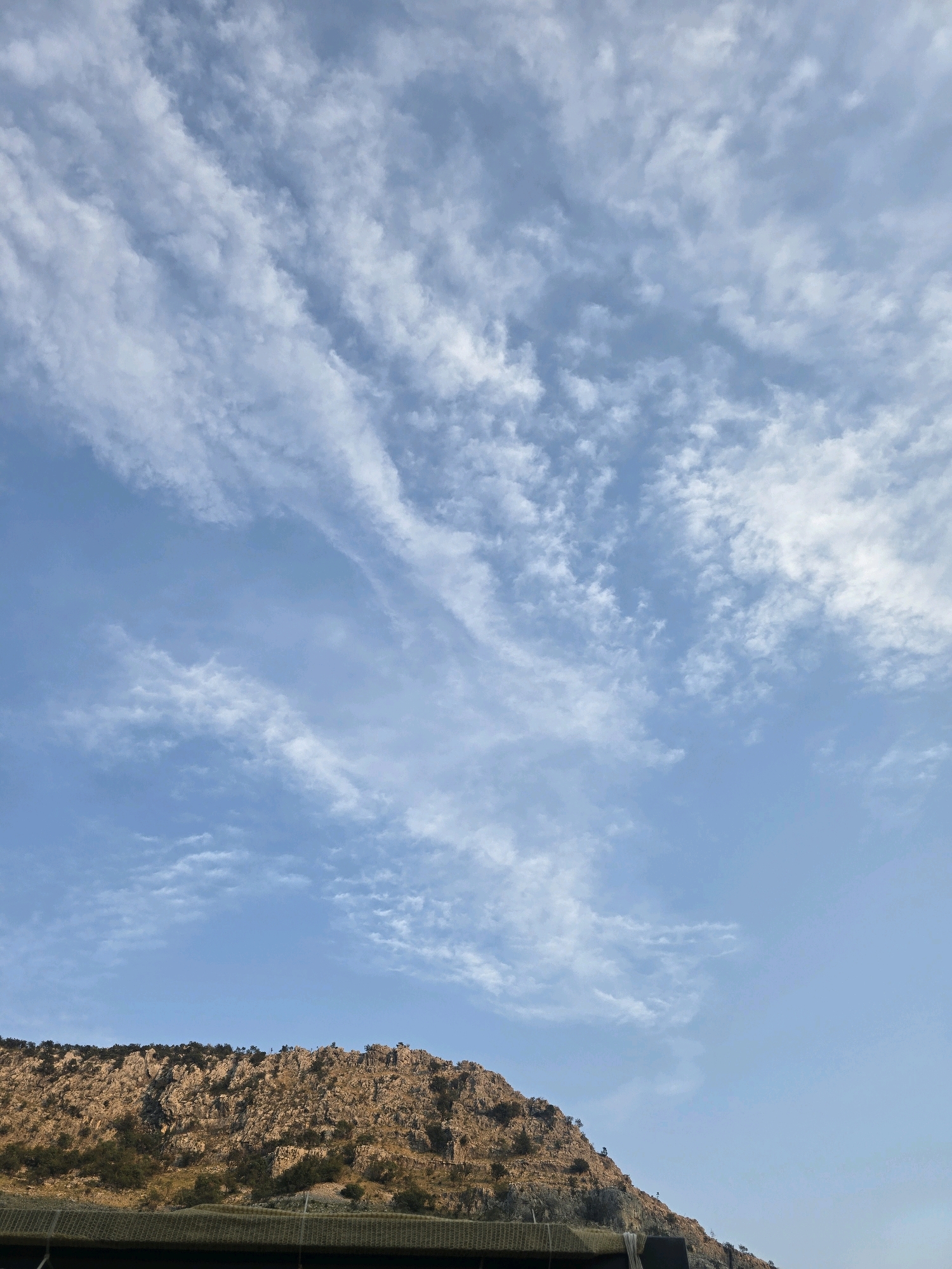 The image shows a landscape scene with a rocky mountainside in the foreground and a sky filled with wispy clouds in the background. The mountain is covered in sparse vegetation and appears to be illuminated by sunlight, casting shadows. A dark, blurred structure is visible at the bottom of the frame, possibly a fence or netting. The sky is a clear blue with streaks of white clouds, suggesting a partly cloudy day. The scene is likely outdoors, possibly in a mountainous region near Podgorica, Montenegro, given the geological features. There are no people or animals visible in the image.