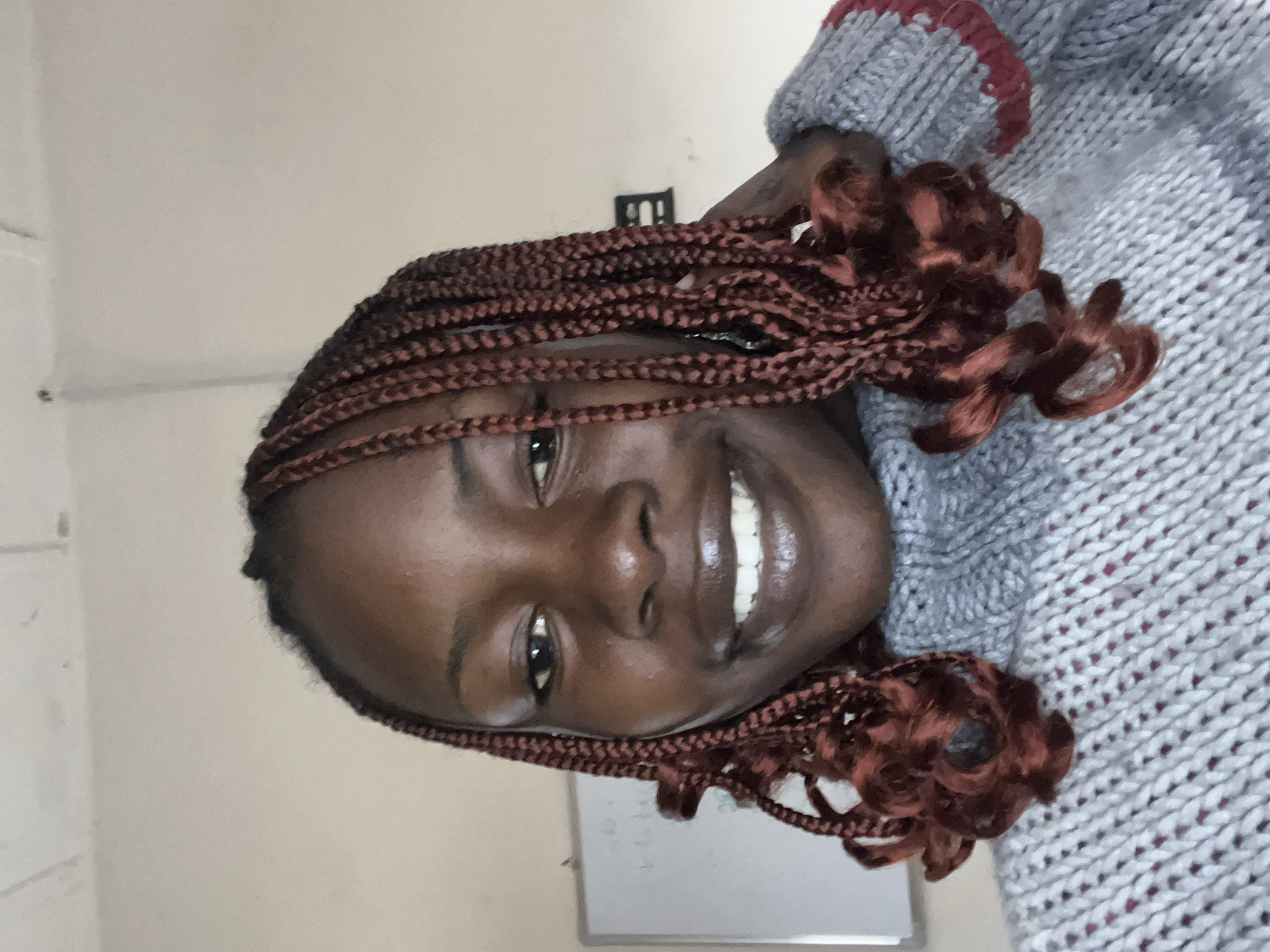 A young Black woman with braided hair, possibly dyed a reddish-brown color, smiles at the camera. She is wearing a gray turtleneck sweater with red trim. The background appears to be an indoor environment with a plain wall and a whiteboard visible on the left. The lighting is soft and diffused. The image is a close-up shot, focusing on the woman's face and upper body. There is no specific indication of the location being Jos, Nigeria, within the image itself.