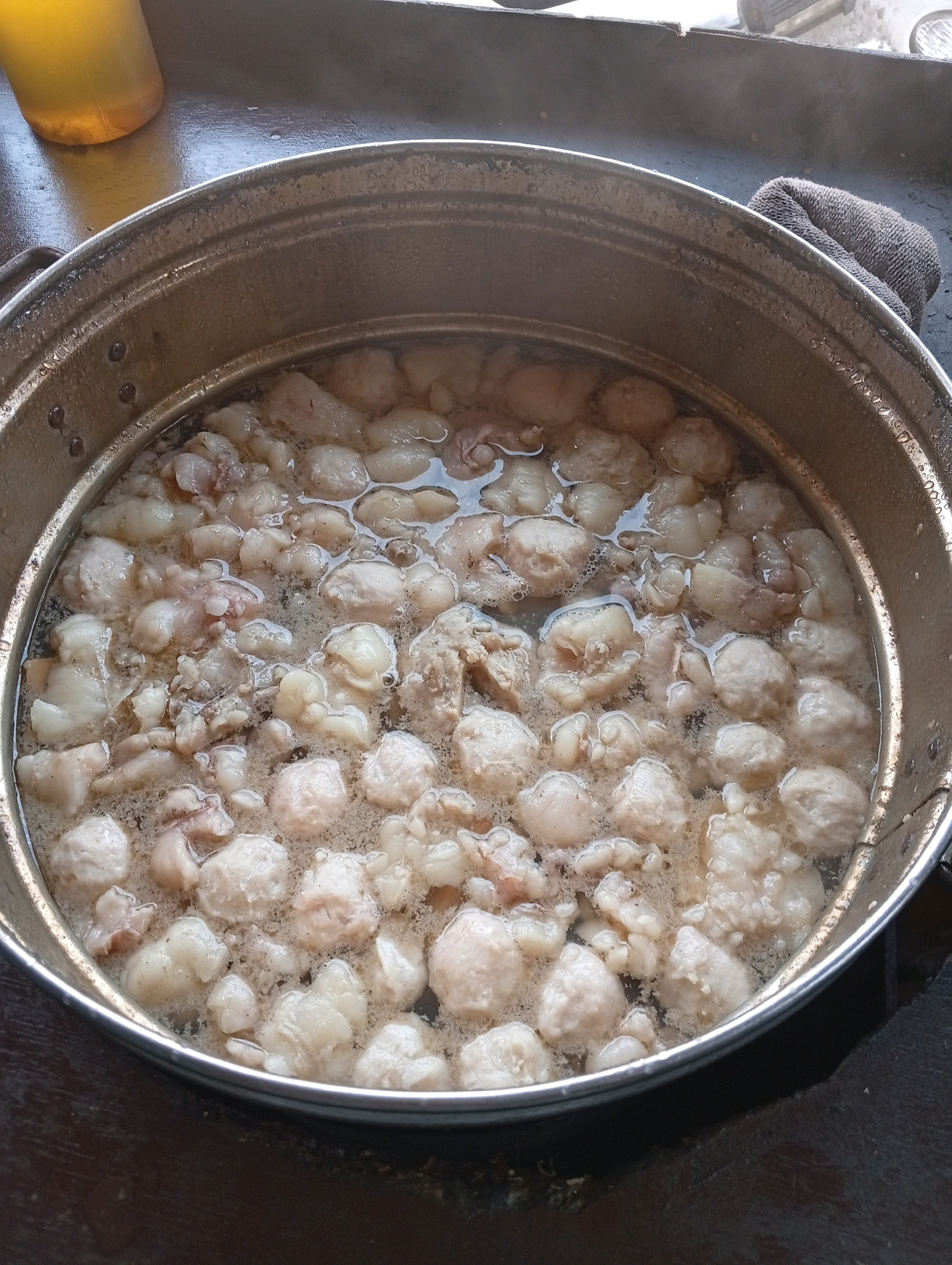 A high-angle, close-up shot shows a stainless steel pot filled with small, pale, spherical meatballs and pieces of what appears to be animal fat or cartilage, simmering in a light-colored broth. The pot is placed on a dark, wooden surface. To the upper left of the pot, a portion of a yellow plastic container with liquid is visible. A dark gray cloth is draped over the right edge of the pot. The environment suggests a casual, possibly outdoor or semi-outdoor food stall or kitchen setting in East Jakarta, Indonesia.