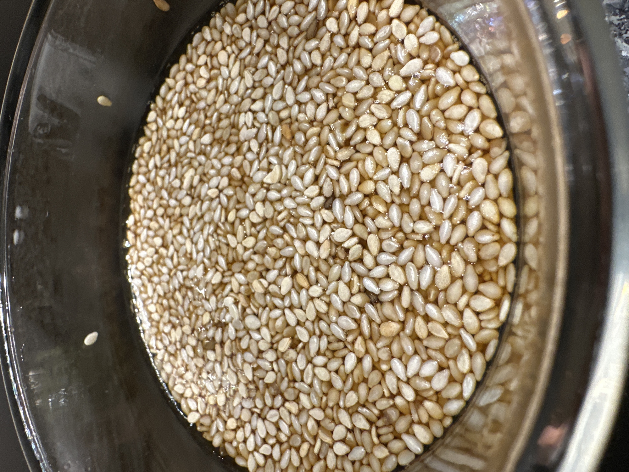 The image depicts a close-up, overhead shot of a clear, circular container filled with sesame seeds submerged in a light-colored liquid, potentially water or brine. The container appears to be made of glass or a clear plastic material. The seeds are densely packed, covering the entire visible area within the container. The overall scene presents a uniform texture dominated by the shape and color of the seeds, which are a pale tan hue. The environment is not discernible, and there are no people or actions captured in the frame. The city of Mexico City, Mexico, is not visible or suggested by the elements within the image.
