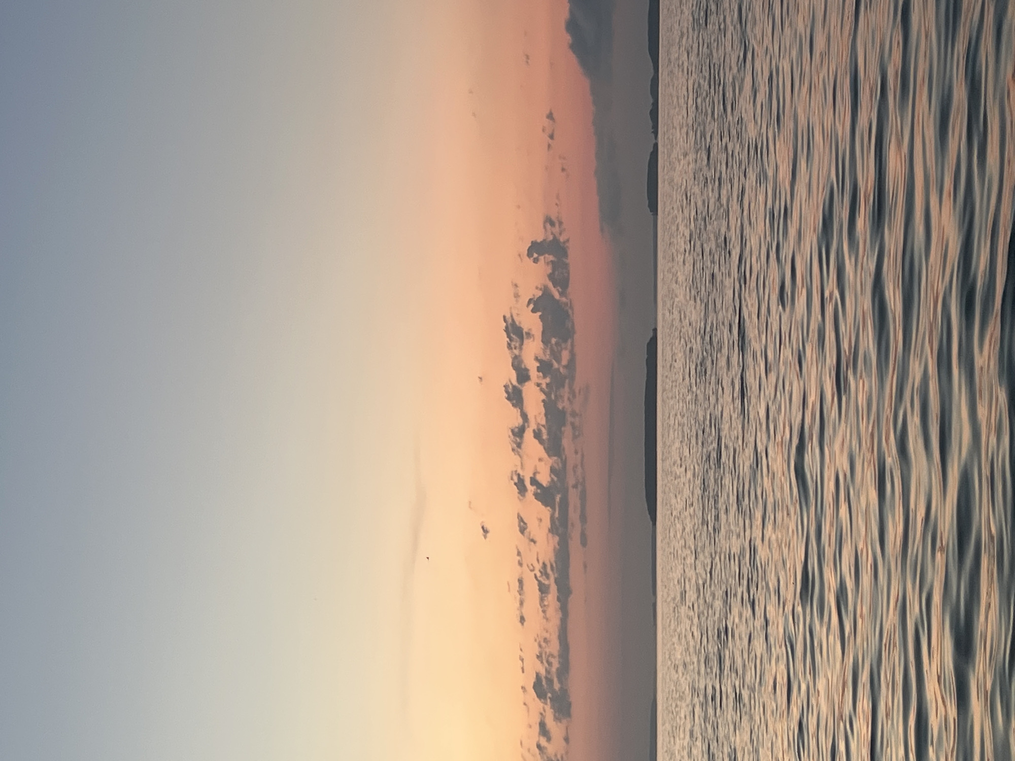 A serene body of water, likely a lake or calm sea, stretches across the foreground of the image. The water's surface is gently rippled, reflecting the soft hues of the sky. In the distance, a thin, dark landmass, possibly islands or a coastline, is visible along the horizon. Above the water, the sky displays a gradient of colors, transitioning from a pale blue at the top to warm oranges and pinks near the horizon, suggesting it is either sunrise or sunset. A scattering of small, dark clouds dots the sky. The scene is devoid of any people, structures, or discernible activity, presenting a natural and tranquil landscape. The location is specified as Novopichugovo, Russia.