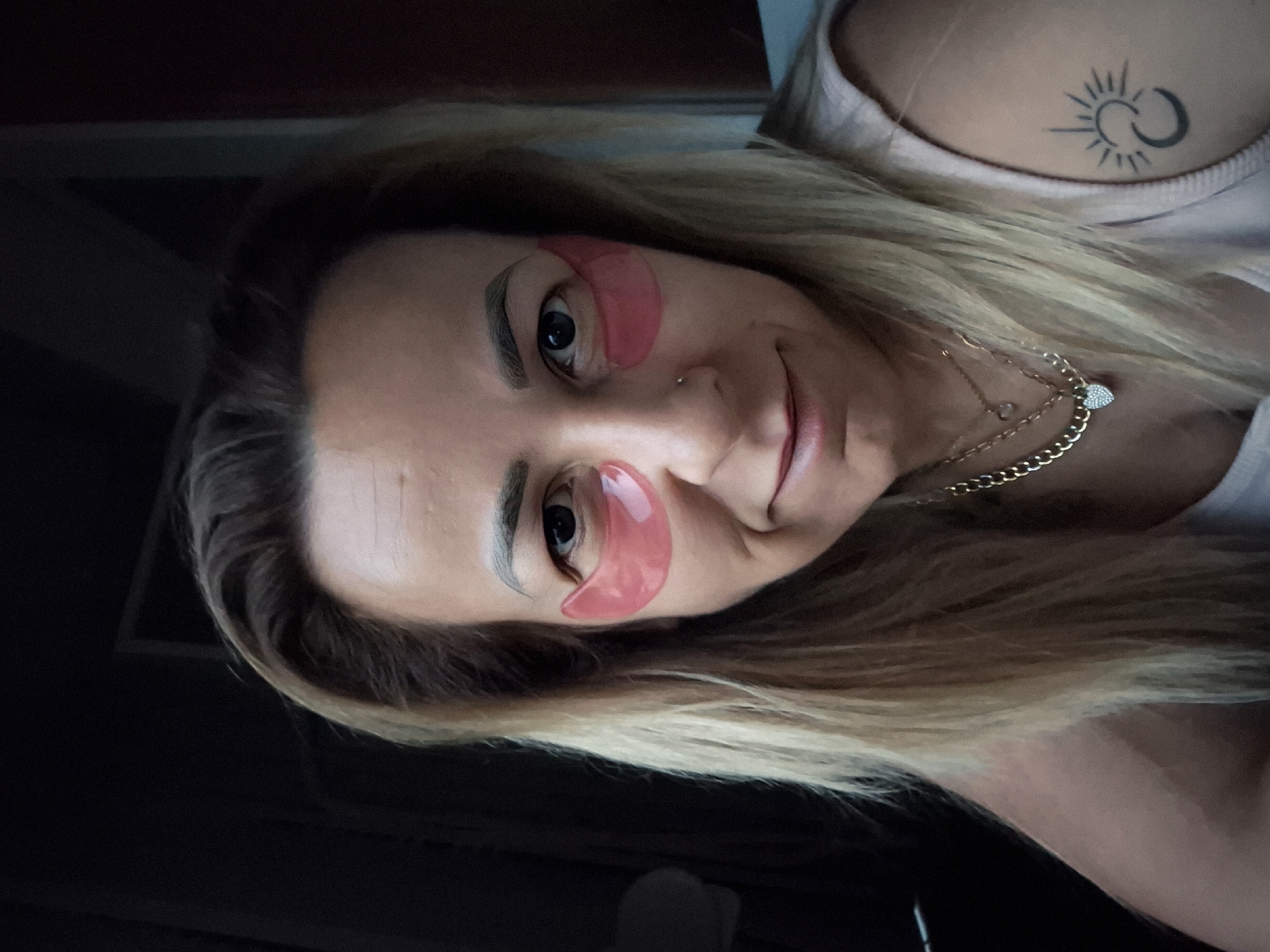A medium shot photograph depicts a woman with blonde hair, wearing pink under-eye patches and a light pink top. She has a nose ring, multiple necklaces, and a tattoo of a crescent moon and sun on her right shoulder. The background is dark, with a hint of a brown wall on the right side.