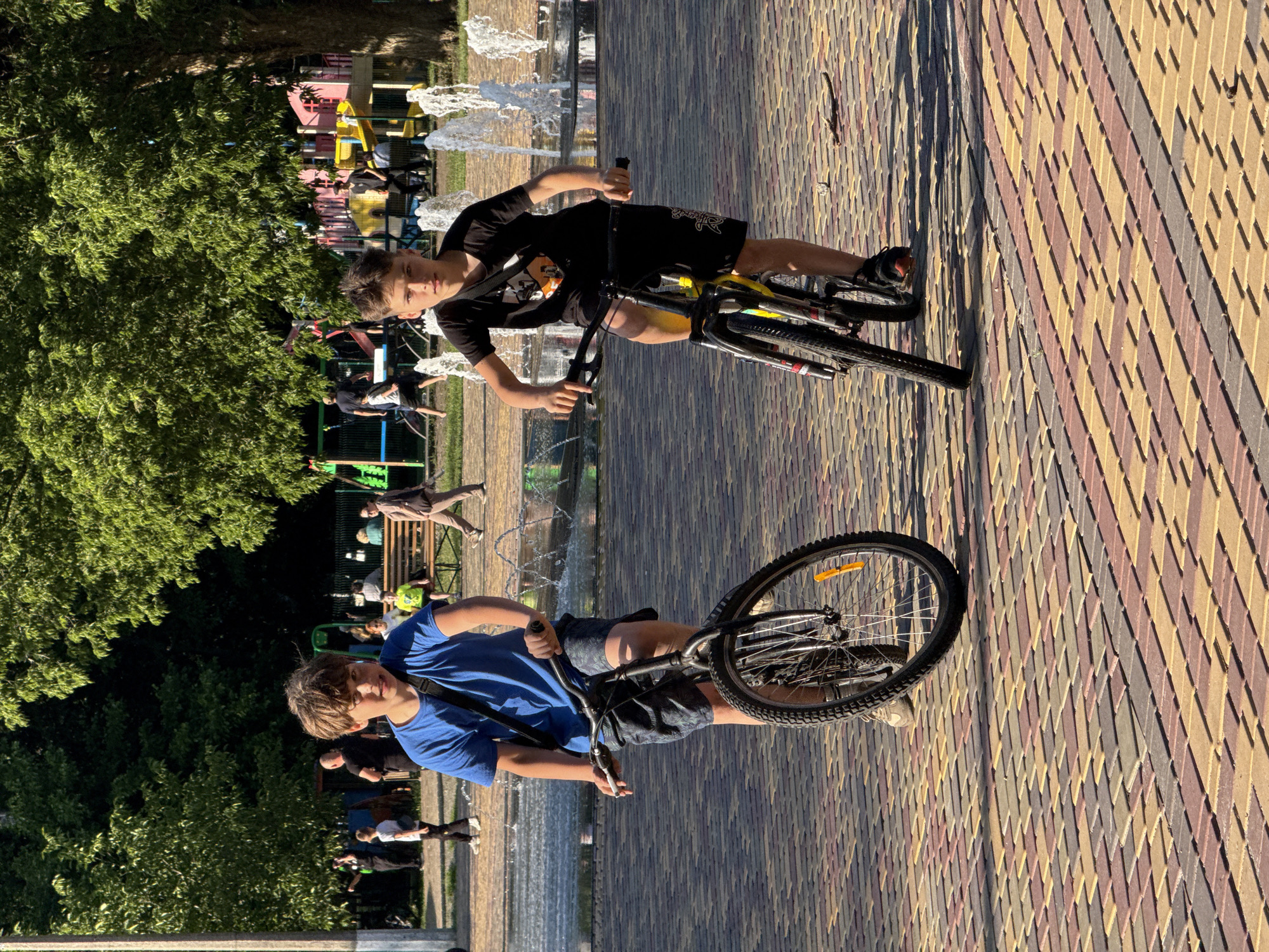 Two young boys ride bicycles in a park. The boy on the left wears a blue t-shirt and patterned shorts. The boy on the right wears a black t-shirt and black shorts. Both boys are looking at the camera. They are on a brick-paved path. In the background, there is a playground with colorful structures and a fountain. Trees with green foliage are visible behind the playground. The sun is shining, casting shadows on the ground.