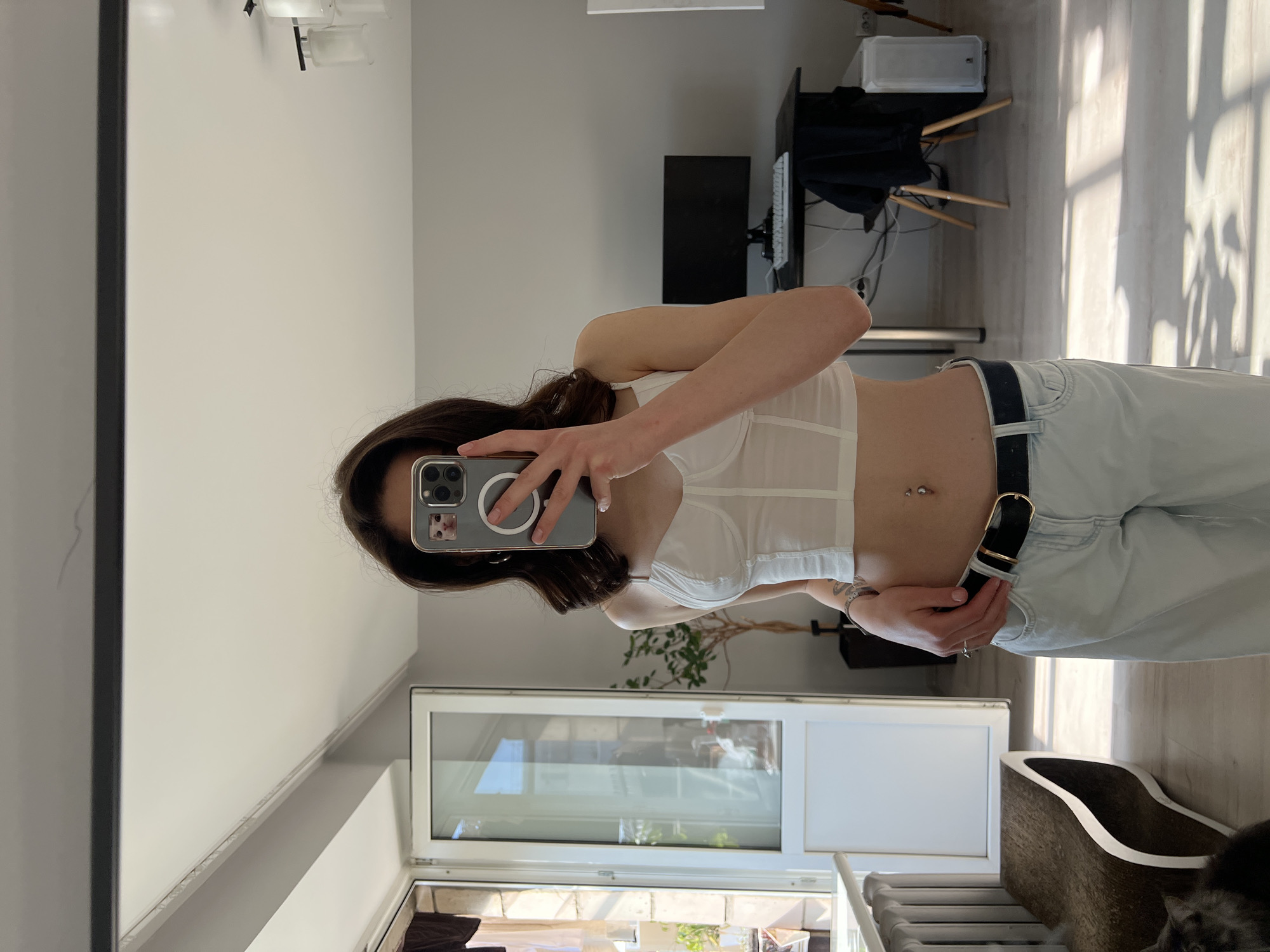 A young woman is standing indoors, taking a mirror selfie. She is wearing a white cropped tank top and light blue wide-leg jeans, accessorized with a black belt and a navel piercing. Her dark hair is styled in loose waves, and she is holding a smartphone with a cat sticker on its case. The background shows a room with a large window, a desk with a computer monitor and keyboard, and a potted plant. There is also a radiator and a curved cat scratching post visible. The lighting suggests it is daytime, with sunlight streaming through the window and casting shadows on the floor. The scene appears to be an apartment. The city of Ternopil, Ukraine is not discernible from the image.