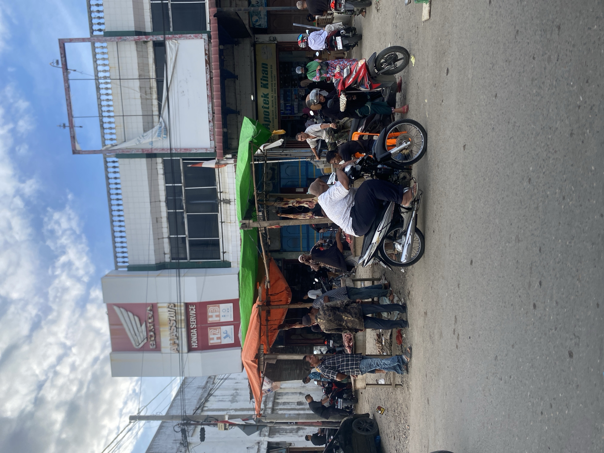 The image depicts a street scene in Pantegajah, Indonesia. The focus is on a row of businesses along the roadside, with a notable Honda service sign prominently displayed. In front of these businesses, a group of men are gathered. One man is sitting on a motorcycle, while others are standing or conversing. Several motorcycles are parked nearby. Overhead awnings are visible, providing shade. The street shows signs of typical urban activity. The sky is cloudy, with sunlight partially illuminating the scene. Visible text includes "Apotek Khan" with an address in Matang Glumpang Dua.