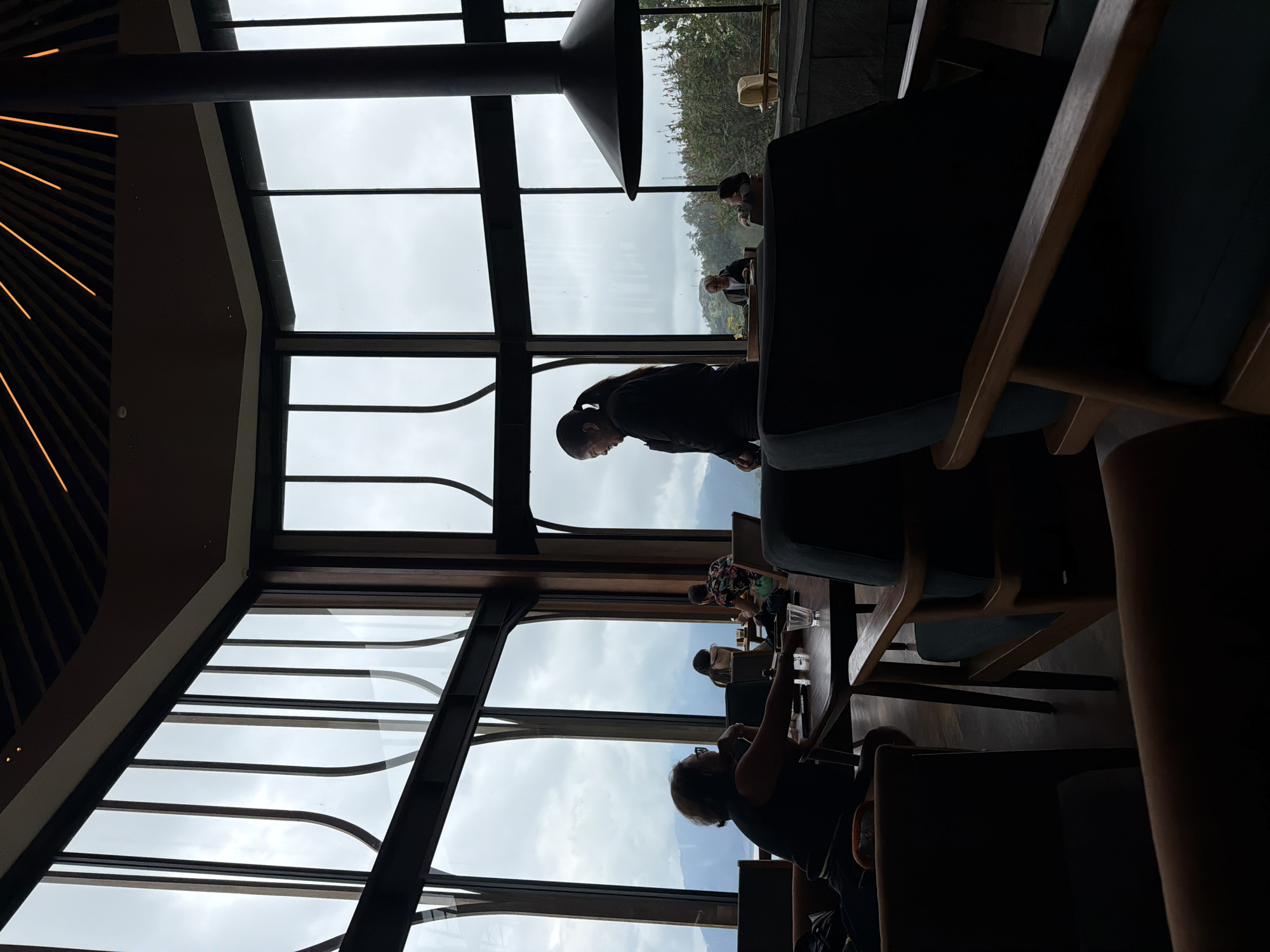 This image depicts the interior of a modern establishment in Kintamani, Indonesia, characterized by expansive windows offering a view of misty mountains and lush greenery. Several individuals are seated at tables, appearing to be engaged in conversation or enjoying the scenery. One woman, silhouetted against the large windows, is standing and seems to be interacting with someone seated at a table. The furniture consists of dark, upholstered chairs and wooden tables, suggesting a comfortable and contemporary setting. A large, black chimney hood dominates the right side of the frame, adding a striking architectural element to the space. The overall ambiance is one of relaxed sophistication, with the natural landscape serving as a prominent backdrop.