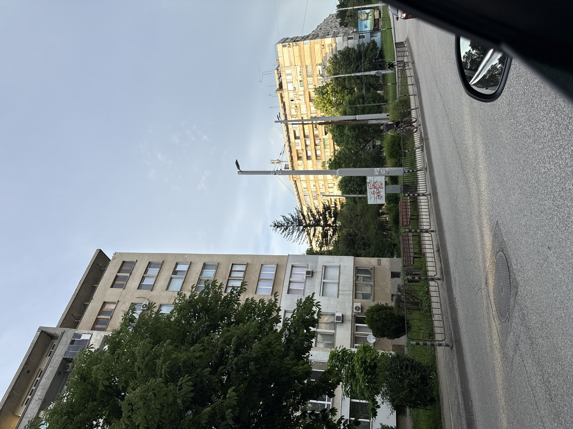 The image depicts a street scene in Pleven, Bulgaria. On either side of the road are buildings, with one being a multi-story structure with multiple windows and at least one air conditioning unit. The other building is also multi-story and appears to be of a similar construction style. Trees are visible in front of both buildings, partially obscuring the lower floors. A lamppost stands beside the road. Metal barriers line the sidewalk next to the street. People on bicycles can be seen along the road. A car's side mirror is visible in the top right corner of the image, indicating the photo was taken from inside a vehicle. The sky is overcast.