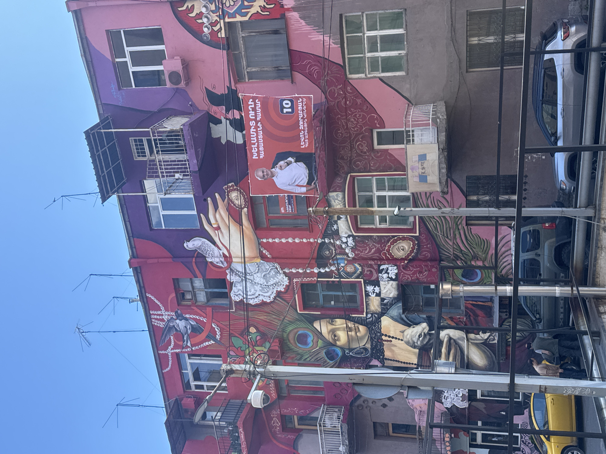 Here is a detailed description of the image:
The image depicts a mural on the side of a building in Yerevan, Armenia. The building is a multi-story structure with various windows, balconies, and architectural features. The mural covers much of the building's facade, featuring a colorful and eclectic mix of artistic elements.
The mural includes images of faces, hands, birds, peacock feathers, jewelry, and abstract patterns. The color palette is vibrant and includes shades of pink, purple, red, and green.
Affixed to the building, within the mural area, is a political campaign poster featuring a man in a white shirt and the text "ԼԵՎՈՆ ԶՈՒՐԱԲՅԱՆ" (Levon Zurabyan) and "10".
The area in front of the building appears to have a mix of utility poles, wires, and building supports. Parked cars are visible on the right side of the building.
The sky above the building is a clear blue.