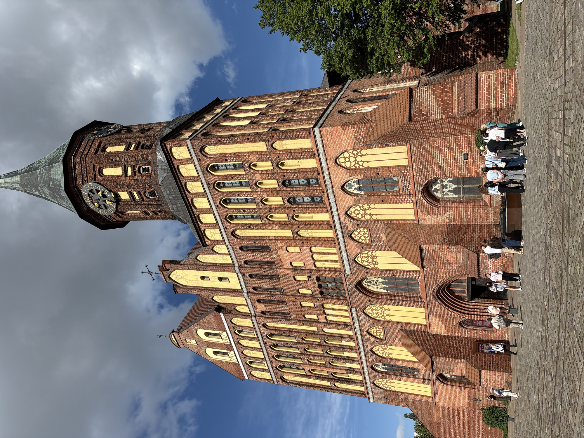 The image captures a grand, historic cathedral, identified as the Kaliningrad Cathedral (Königsberg Cathedral), under a partly cloudy sky. The structure is predominantly built of reddish-brown brick, featuring intricate Gothic architectural details.
The cathedral's most prominent feature is its tall, pointed spire, topped with a dark, possibly copper, roof. Below the spire, a large clock face with gold Roman numerals is visible, indicating a time around 10:10. The building's facade is adorned with numerous windows, many showcasing delicate yellowish-gold tracery and pointed arches, characteristic of Gothic design. Some windows are tall and narrow, while others are round or have simpler arched forms. The structure is supported by visible buttresses, adding to its imposing silhouette.
Several groups of people are scattered on the paved forecourt in front of the cathedral. They appear to be visitors or tourists, dressed in casual clothing, observing the building or gathering together. Their presence suggests a public or historical site popular for sightseeing.
The setting is outdoors, bathed in bright daylight, indicating a clear day despite the presence of white and grey clouds in the sky. Patches of vivid blue sky are also visible. A lush green tree is positioned towards the upper right of the frame (when upright), providing a natural element against the brickwork. The ground is covered with a patterned paving, likely cobblestones or similar, extending into the foreground. The scene conveys a sense of historical significance and active tourism.