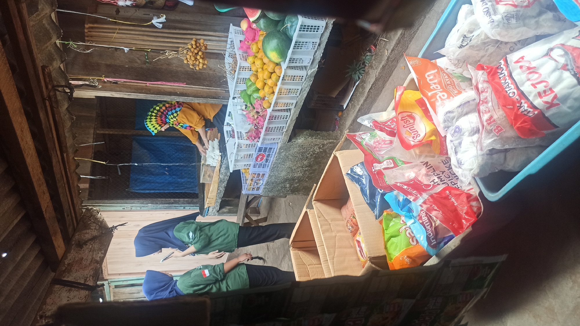 This image depicts a market stall in Trisinar, Indonesia, likely in a rural or semi-urban setting. The stall appears to be an outdoor or semi-enclosed structure with a wooden roof. In the foreground, there are several cardboard boxes and blue plastic bins filled with packaged food items, predominantly plastic bags containing what appear to be frozen or dried food products such as meatballs and snacks. Some of the packaging has Indonesian text visible, like "Indomimi," "Sunfish," and "Kejora."
In the middle ground, a woman wearing an orange shirt and a colorful headscarf is seen behind a counter, actively sorting or preparing some items. To the left, two young women wearing hijabs and green jackets with an Indonesian flag emblem are standing and looking at a smartphone.
The background shows more of the market structure, including wooden planks and a blue tarp. A shelf behind the woman in the orange shirt displays various fruits in white plastic crates, including watermelons, dragon fruit, oranges, and what appear to be longans hanging from a string. The overall impression is a bustling local market scene.