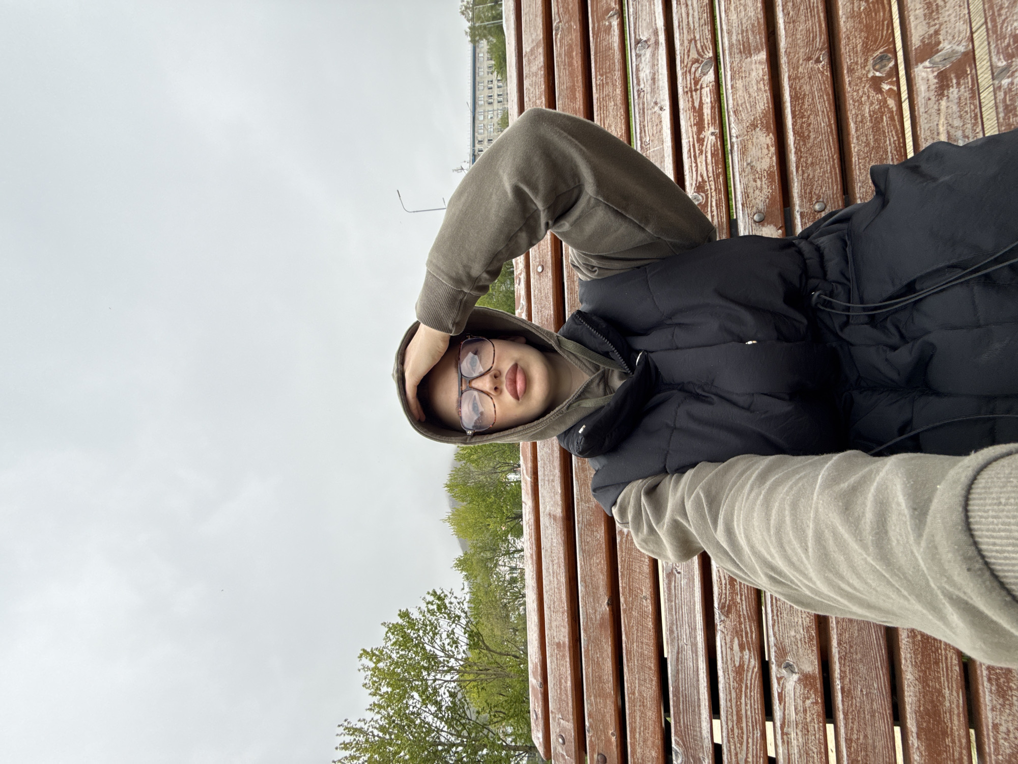A young person with fair skin, brown eyes, and brown lips is sitting on a wooden park bench, holding their right hand to their forehead. They are wearing a dark puffer vest over a brown hooded sweatshirt and large-rimmed glasses. The bench is made of weathered wooden planks. In the background, there are trees with green foliage, a cloudy sky, and a building with multiple windows. The scene appears to be in a park or public outdoor space, potentially in the city of Petropavlovsk-Kamchatsky, Russia, given the environmental context. The overall lighting is diffused and overcast.