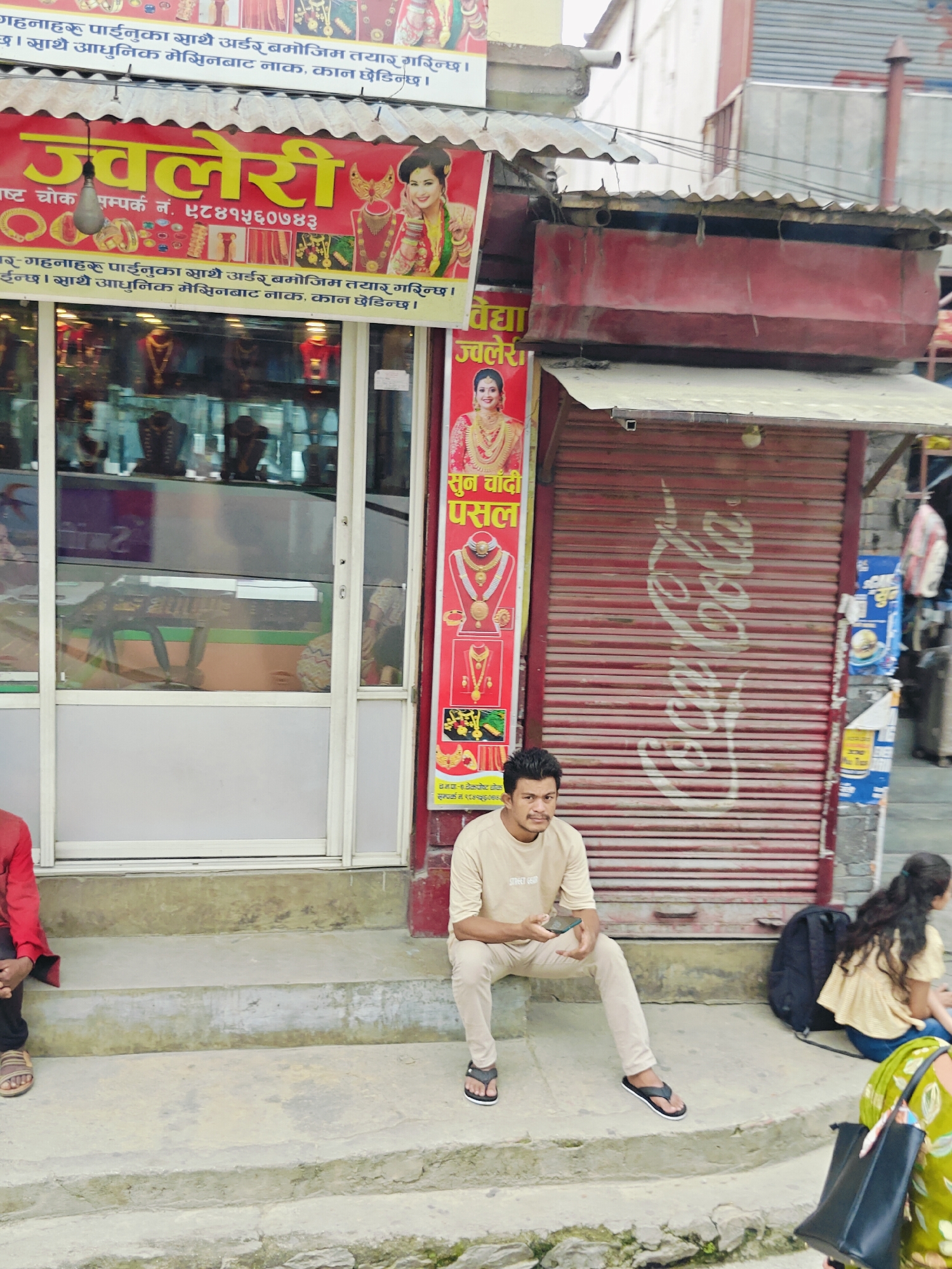 In Chandragiri, Nepal, a street scene unfolds with a jewelry store as the central focus. The store's prominent red sign, featuring the word "Jewelery" in yellow, is adorned with images of women in traditional attire and gold ornaments. To the right of the store, a red metal shutter is partially visible, bearing a faded "Coca-Cola" logo.
Seated on the concrete steps in front of the shutter is a man wearing a beige t-shirt and light-colored trousers. He is holding a mobile phone and looking towards the camera. To his right, another person, partially visible, is seated with a backpack nearby. Further to the right, another individual, wearing a green patterned outfit, is also partially visible. A person in a red top and dark trousers is seated to the left of the man with the phone. The storefront itself features glass display cases filled with various jewelry items. The overall environment appears to be an urban street, with elements of commerce and pedestrian activity.