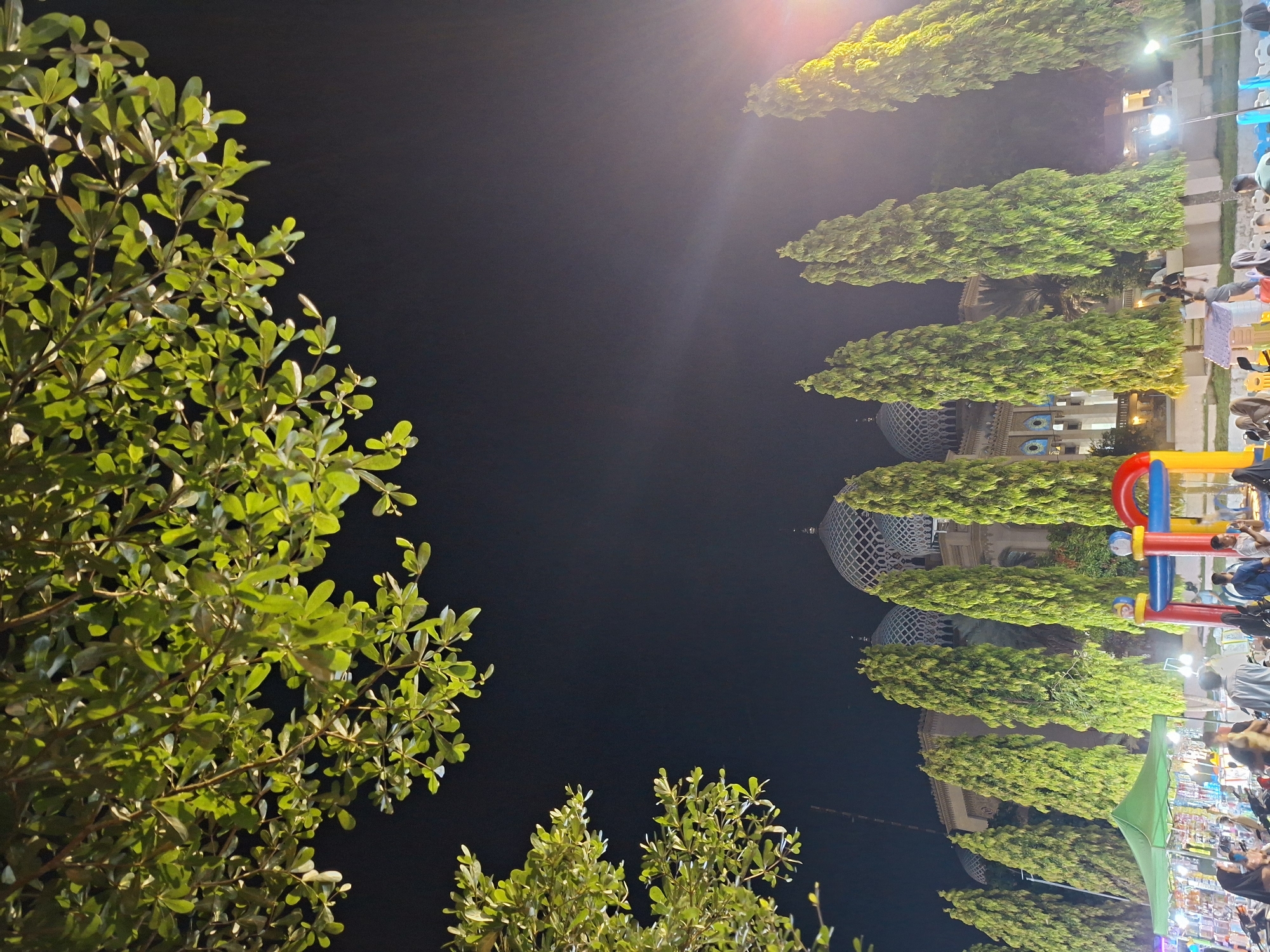 The image depicts a nighttime scene in Lhokseumawe, Indonesia. The view is framed by green foliage of trees on the left and at the bottom. The foreground is dark, suggesting it is taken from a lower vantage point. In the middle ground, a line of trimmed green trees leads towards a building with distinctive blue domes and architectural details suggesting a mosque or religious building. People can be seen in the right portion of the image, with hints of a playground structure. The lighting is artificial and comes from streetlights and building illumination, casting bright highlights and shadows. The sky is dark, and the overall atmosphere suggests a public space visited at night.