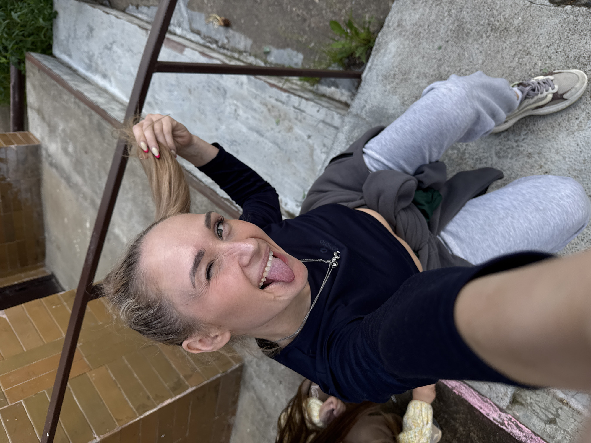 The main subject of the image is a young woman taking a playful selfie from an overhead angle. She is winking with her right eye, sticking out her tongue, and holding a section of her blonde hair. Her nails are manicured with white tips and a red detail near the cuticles. She wears a dark long-sleeved top with the text "OMOTO" visible on the chest, light grey sweatpants, and light-colored sneakers with brown accents. A silver necklace with small charms is visible around her neck.
The setting appears to be an outdoor staircase in Chaikovsky, Russia. The stairs are multi-level, with a metal handrail running alongside. The upper parts of the staircase are made of grey concrete, while the lower steps are covered in yellowish-brown tiles arranged in a herringbone pattern. Some natural elements, including green plants and weeds, are visible around the concrete steps, suggesting an urban or semi-urban outdoor space. A partial view of a second person with brown hair and a light-colored top is visible at the very bottom of the frame. There's a faint reddish-pink paint stripe on a concrete ledge in the bottom right.
The overall mood of the image is lighthearted and energetic. It appears to be daytime, likely under an overcast sky, given the diffused lighting.