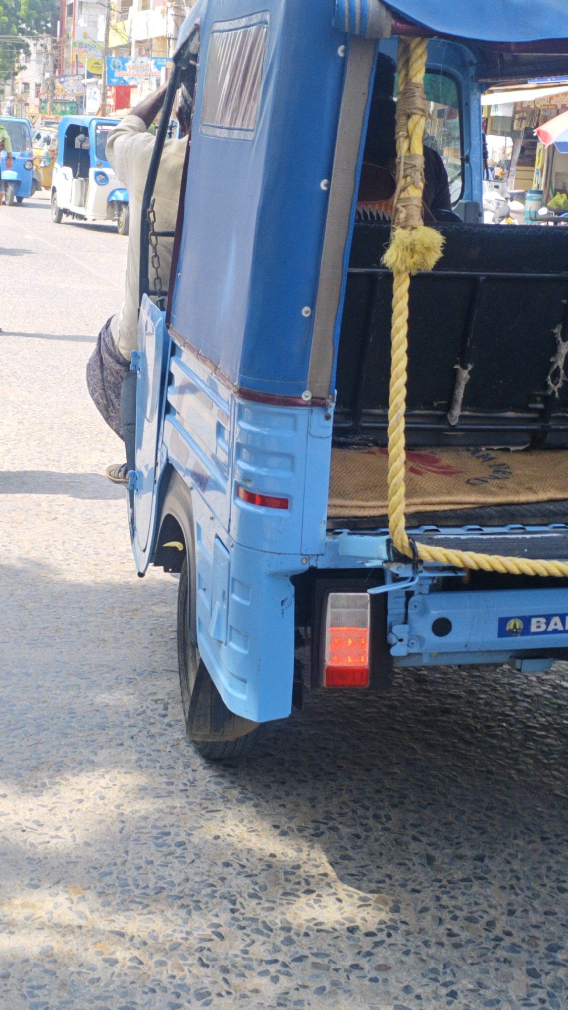 The image shows a blue auto-rickshaw, a common mode of transport in India, parked on a paved street. The auto-rickshaw has a canvas roof and is partially open at the back, revealing a woven mat inside. A yellow rope is tied to the side of the auto-rickshaw. In the background, there are buildings and other auto-rickshaws, suggesting an urban environment. The scene appears to be a street in Pamur, India.