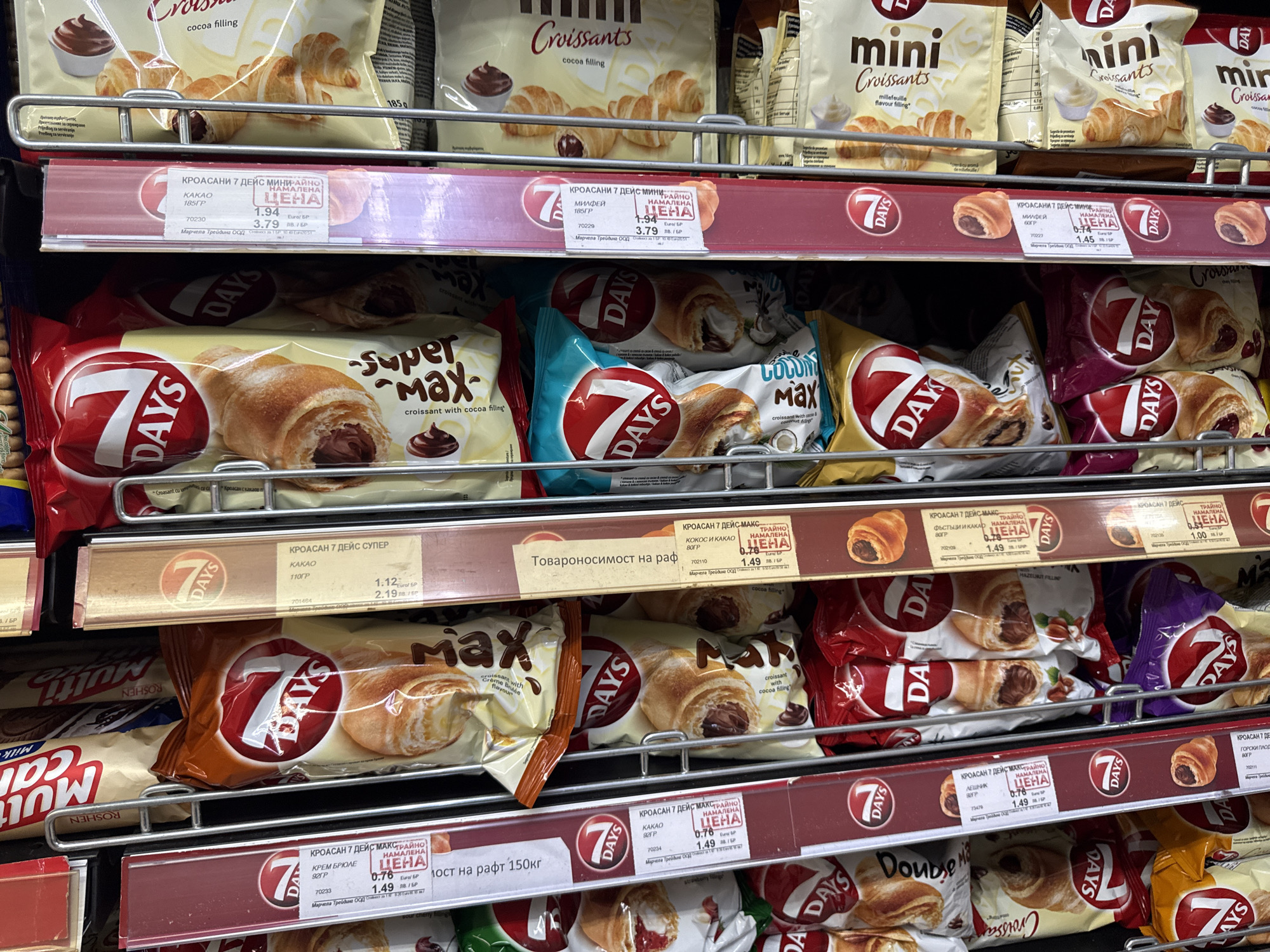 The image depicts a grocery store shelf stocked with various flavors of "7 Days" croissants. The croissants are packaged in individual bags and arranged in rows, with price tags displayed in front of each row. The price tags are in Bulgarian currency (лв) and display both the discounted price and the regular price. Different croissant flavors are visible, including cocoa, crème brûlée, coconut and cocoa, hazelnut, and forest fruit. The shelf is made of metal and has a dark red horizontal stripe across the front. The background shows other products on nearby shelves, suggesting this is a general view of a snack aisle in a grocery store.