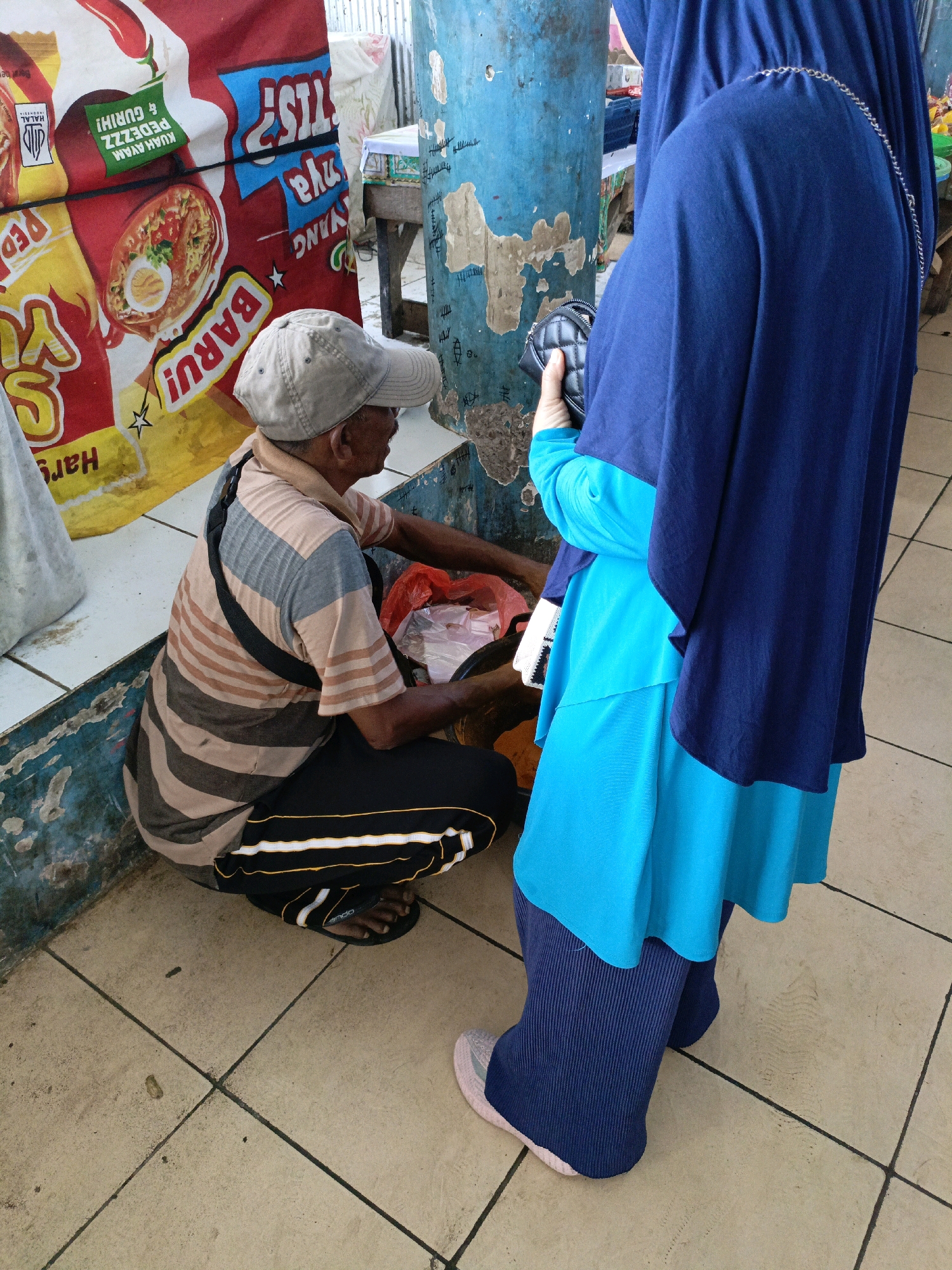 A medium shot captures two individuals in what appears to be an outdoor market stall in the city of Ela-ela, Indonesia. On the left, a man wearing a light brown and gray striped t-shirt, black pants with yellow stripes, a grey baseball cap, and black sandals is crouched down. He is handling items in a red plastic bag and a clear plastic bag, possibly involved in a transaction. To the right, a person wearing a dark blue hijab and a bright turquoise tunic over dark blue pleated pants stands facing the man. They are holding a black quilted purse. The background features a blue, distressed pillar with graffiti, a large red and yellow banner advertising instant noodles with text in Indonesian, and a tiled floor.