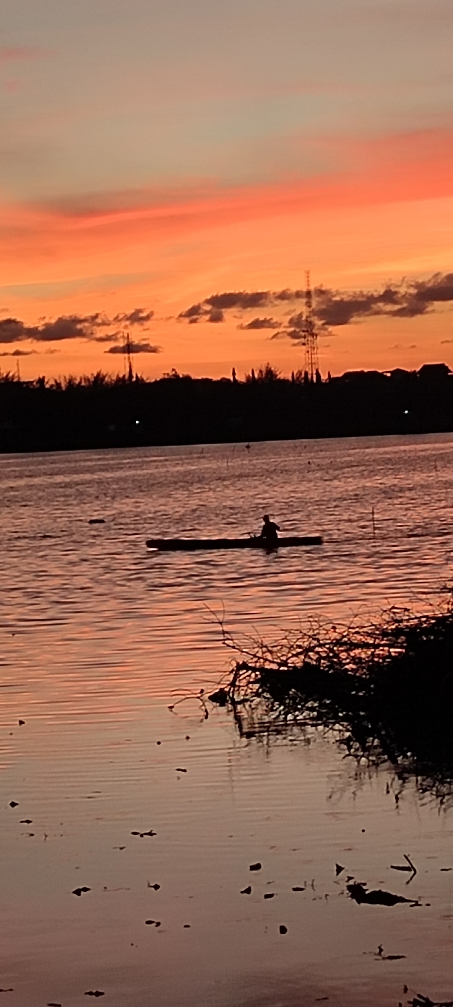 This is a vertical, medium-shot photograph captured at sunset in Lhokseumawe, Indonesia. The scene depicts a tranquil body of water, likely a river or estuary, with a silhouetted figure paddling a long, narrow boat. The sky above is a vibrant canvas of orange and pink hues, streaked with wispy clouds, indicating the sun is low on the horizon.
The silhouette of the person in the boat is small, emphasizing the vastness of the water and the sky. The water itself reflects the colors of the sunset, creating a warm, reddish-pink glow. Scattered on the water's surface are small, dark objects, possibly debris or aquatic plants.
In the distance, a dark silhouette of trees and structures lines the horizon. A tall transmission tower is visible among the trees, a common feature in populated areas. The overall mood of the image is serene and peaceful, evoking a sense of calm as the day draws to a close. The soft lighting and the solitary figure in the boat contribute to a contemplative atmosphere. No visible text is present in the image.