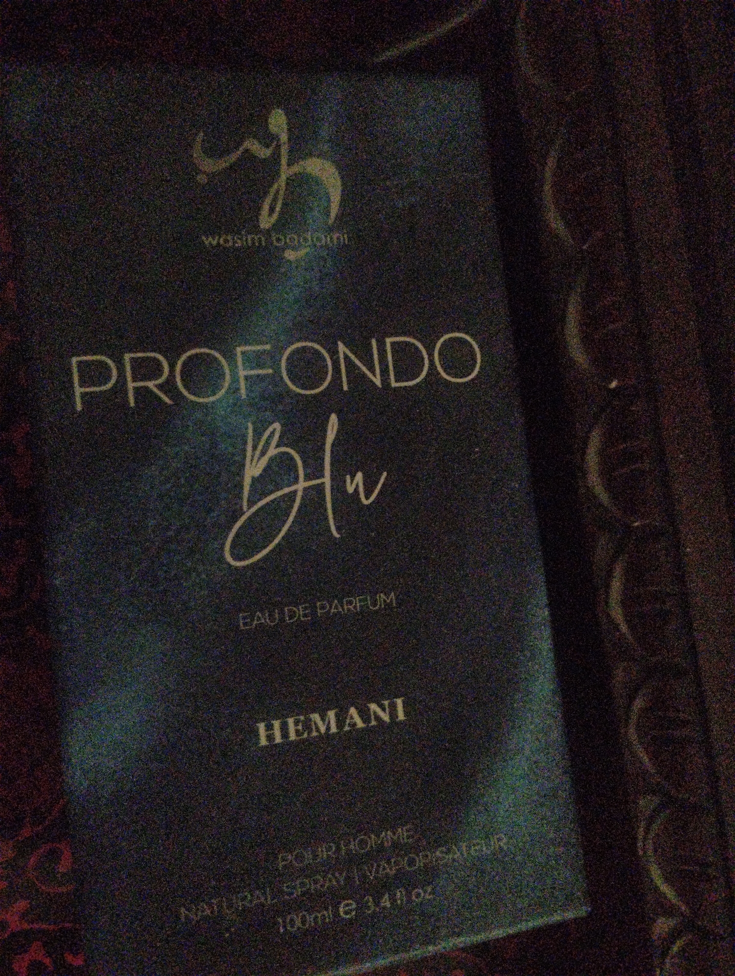 This is a close-up, low-light, grainy photograph of a perfume bottle box. The box is dark blue with white and gold lettering. The prominent text on the box reads "PROFONDO Blu" in large font, with "EAU DE PARFUM" below it. There is also a brand name "HEMANI" written in smaller, white font, and at the bottom, "POUR HOMME NATURAL SPRAY | VAPORISATEUR 100ml e 3.4 fl oz". A stylized gold logo and the name "wasim badami" are visible at the top of the box. The background is dark and indistinct, with what appears to be fabric or textured material. The lighting is dim, and the image quality is affected by noise. No people or specific location are visible, and no activity or event is depicted. The focus is solely on the product packaging.