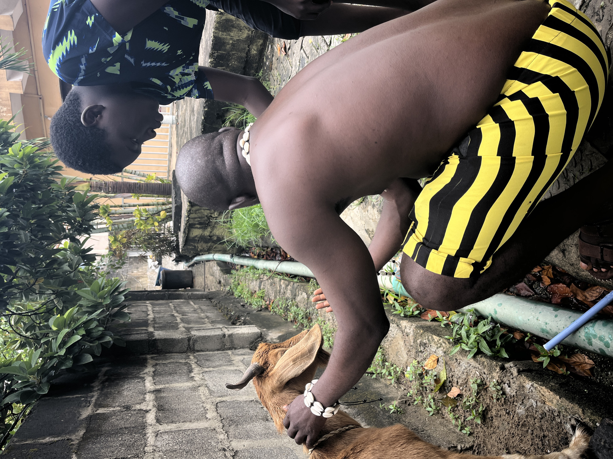 On a paved pathway near Ado, Nigeria, a shirtless dark-skinned man wearing a shell necklace and black and yellow striped shorts holds a brown goat with horns. Another person, possibly a child, in a patterned shirt stands nearby. The setting is a step-way down with green vegetation on the sides. A water hose and a trash can are visible.
