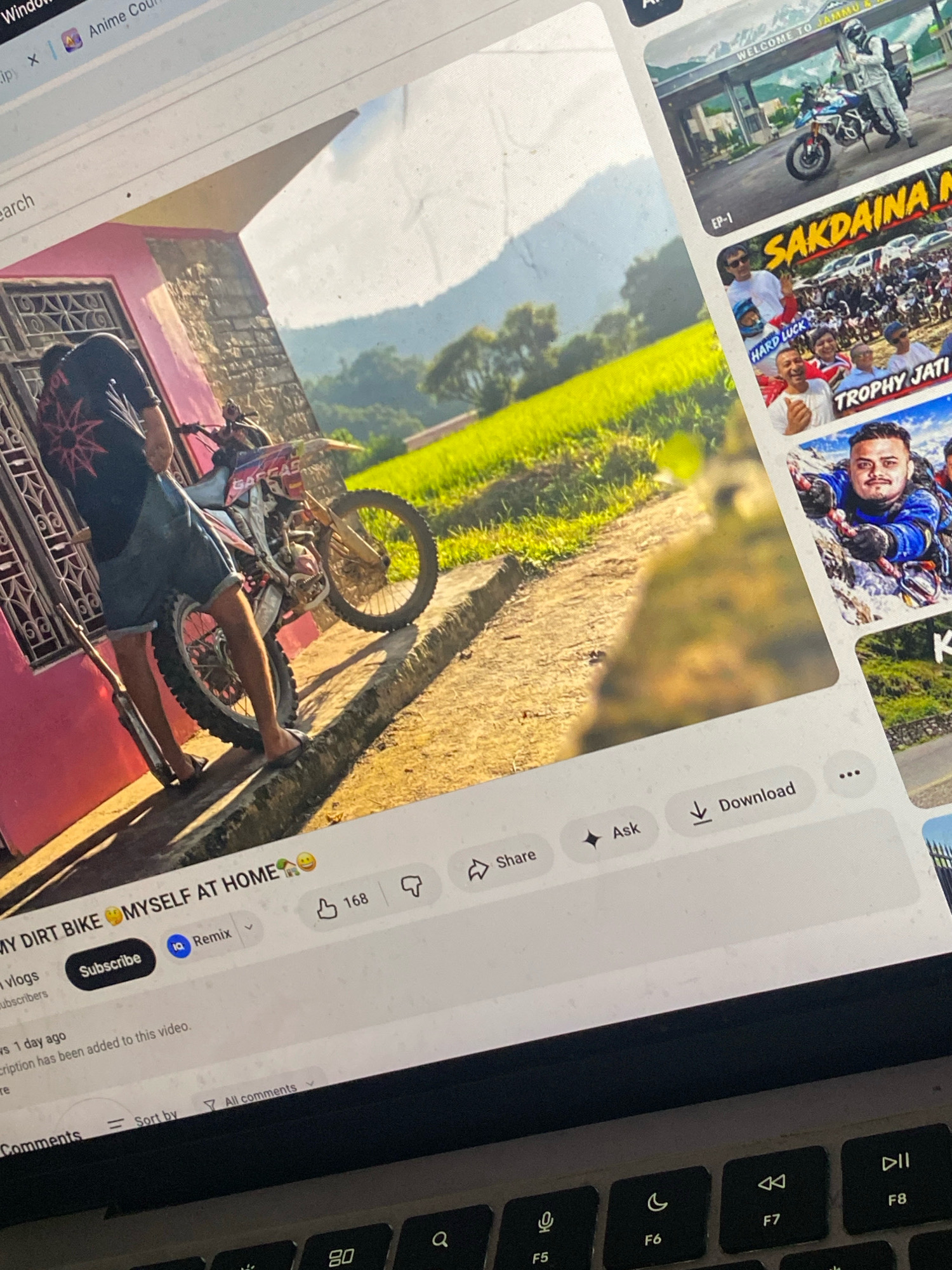 The image displays a laptop screen showing a YouTube video interface with several video thumbnails and playback controls. In the foreground, a young man in a black t-shirt with a red starburst design and denim shorts is standing next to a dirt bike. The bike is positioned on a concrete pathway in front of a pink-colored building with ornate window grills. The background features a vibrant green field with trees and distant mountains under a bright sky. The video title visible is "MY DIRT BIKE MYSELF AT HOME". The YouTube interface also shows options for subscribing to a channel, liking the video (168 likes), commenting, sharing, asking questions, and downloading. The overall scene is a digital representation of online content, likely a vlogging or motorsports video. There is no specific indication of the location being Mahalaxmi, Nepal.
