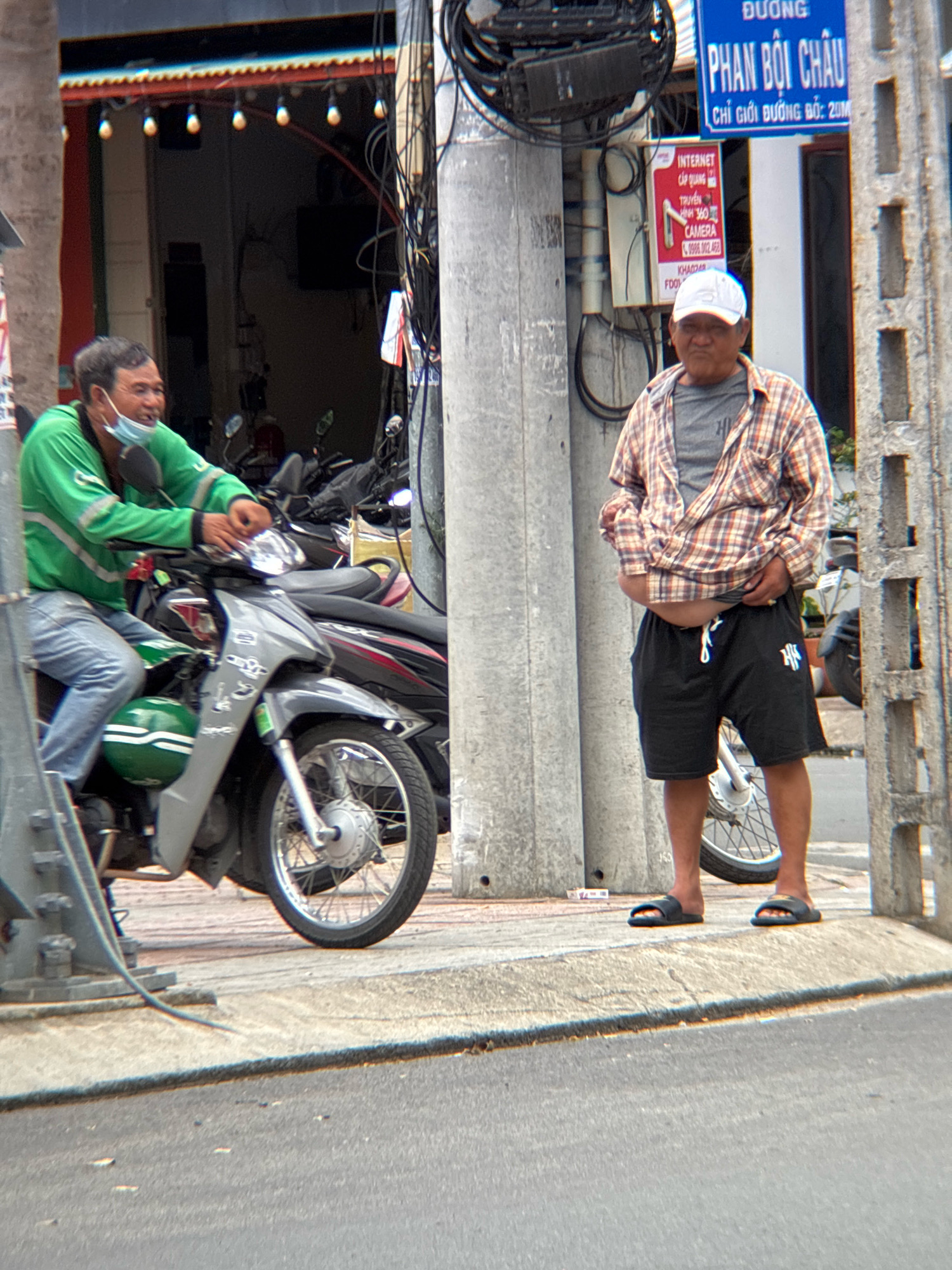 The image depicts a street scene in Nha Trang, Vietnam. In the foreground, a man wearing a plaid shirt, gray t-shirt, and black shorts stands with his hands near his stomach, looking towards the camera. He is wearing black sandals. To the left, another man, wearing a bright green jacket, blue jeans, and a face mask, is seated on a silver motorcycle. He is also looking towards the camera. The motorcycle has a green helmet resting on its side. In the background, a storefront with an awning decorated with lights is visible, along with utility poles and wires. A blue sign with Vietnamese text is mounted on one of the poles, reading "PHAN BOI CHAU." Another red and white sign below it advertises internet and camera services. The ground is paved with concrete and asphalt, with some debris scattered around.