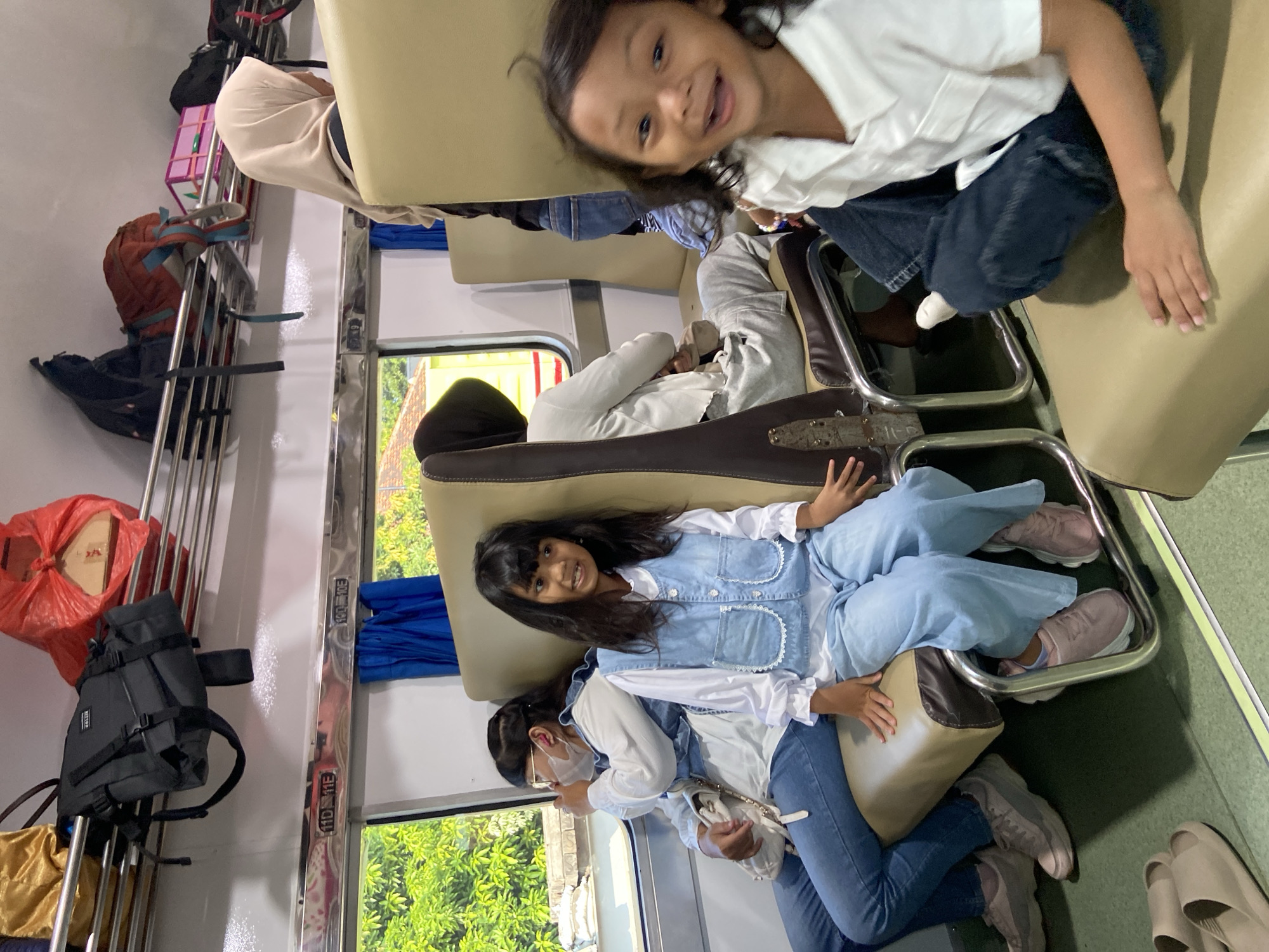 Inside a train carriage, four people are visible. A young child sits in the upper right, a woman and child occupy the middle, and an adult is seated in the background. Luggage is stowed on the overhead rack to the left. The train is moving, as evidenced by the view of greenery outside the window. The location is Probolinggo, Indonesia.