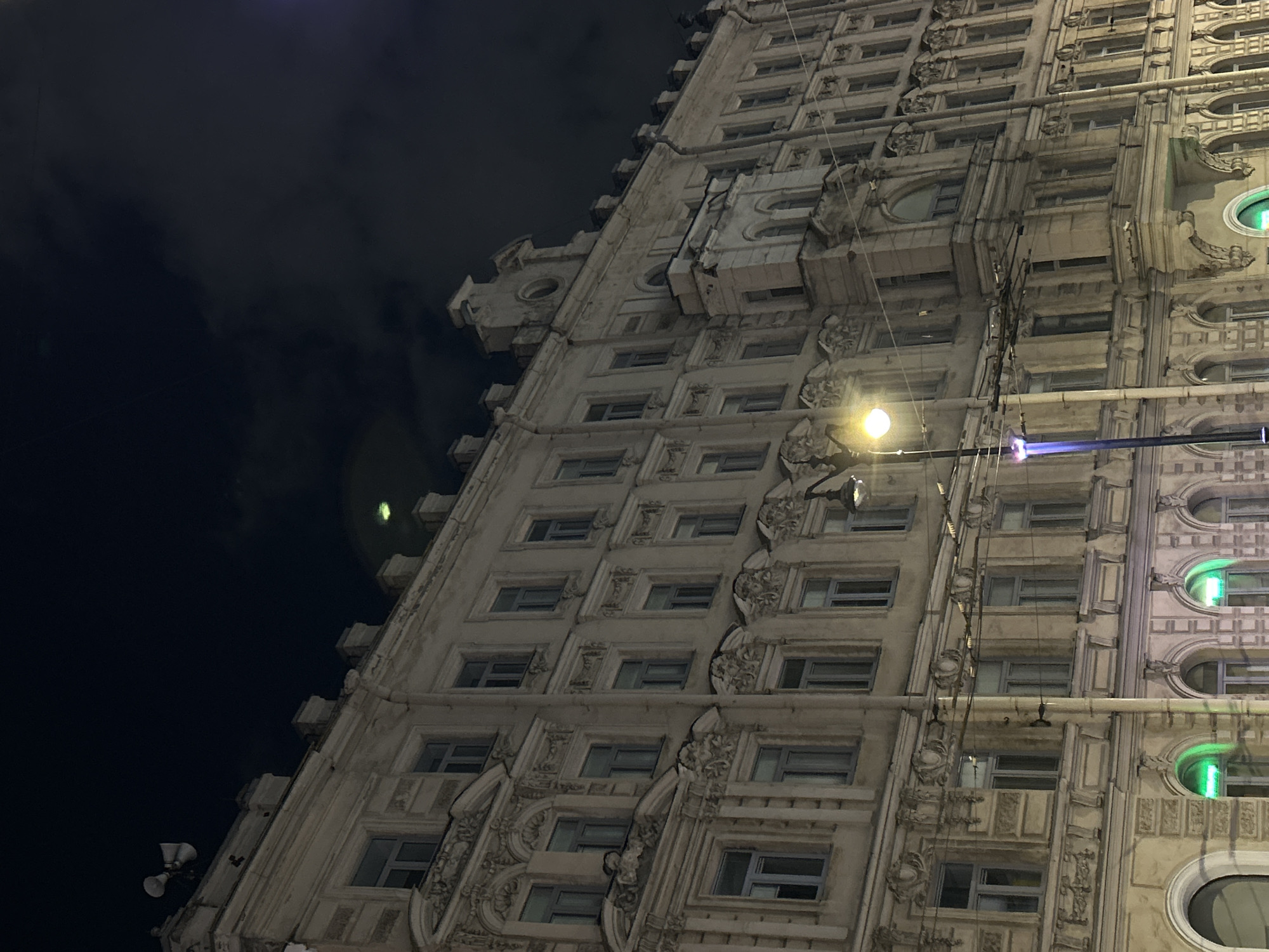 The image displays a grand, multi-story building at night, viewed from a low, upward-tilted angle. The sky is dark, indicating the nighttime setting, with hints of cloud cover and subtle light reflections in the upper left.
The primary subject is the building's highly ornate facade, constructed from a light-colored material, likely stone or stucco, and showcasing intricate architectural details. These include numerous decorative carvings, cornices, pilasters, and elaborate mouldings, suggesting a classical or Baroque revival style consistent with historical architecture found in Saint Petersburg, Russia. The facade features many rows of windows; most are rectangular, but a section on the right side contains distinctive arched and oval-shaped windows.
A bright, circular yellow street lamp is prominent in the mid-right of the frame, casting a warm glow onto the building. Below the lamp, a streak of blue-violet light is visible, likely a lens flare. Several of the oval windows on the far right emit a distinct green light, possibly from interior illumination or decorative fixtures. Various pipes and wires are discernible, running vertically and horizontally across the building's exterior. There are no people visible, allowing the architecture to dominate the scene. The overall impression is one of historical majesty and detailed craftsmanship under the cover of night in an urban environment. No discernible text is visible.