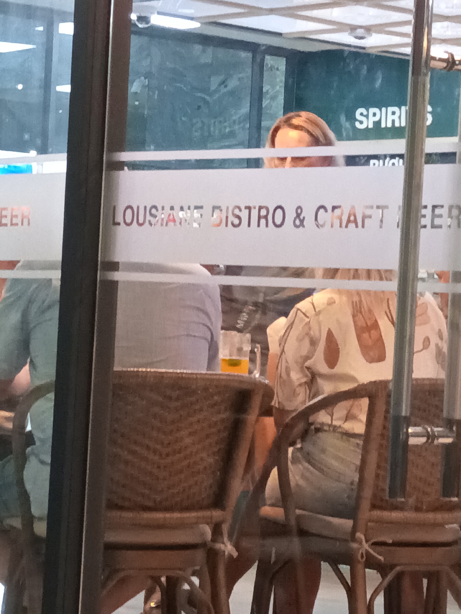 The image shows a storefront window with frosted glass displaying text that reads "LOUSIANE DISTRO & CRAFT BEER". A person with blonde hair is partially visible behind the glass, looking downwards. Inside the establishment, two individuals are seated at tables, one wearing a light blue shirt and the other a patterned shirt with a light blue skirt. A glass of amber liquid, likely beer, is on a table. The scene appears to be a bar or restaurant. The presence of Vietnamese text and signage on other parts of the window suggest the location is in Vietnam. Based on the style of the building and street, it is likely in a tourist area of Nha Trang, Vietnam.