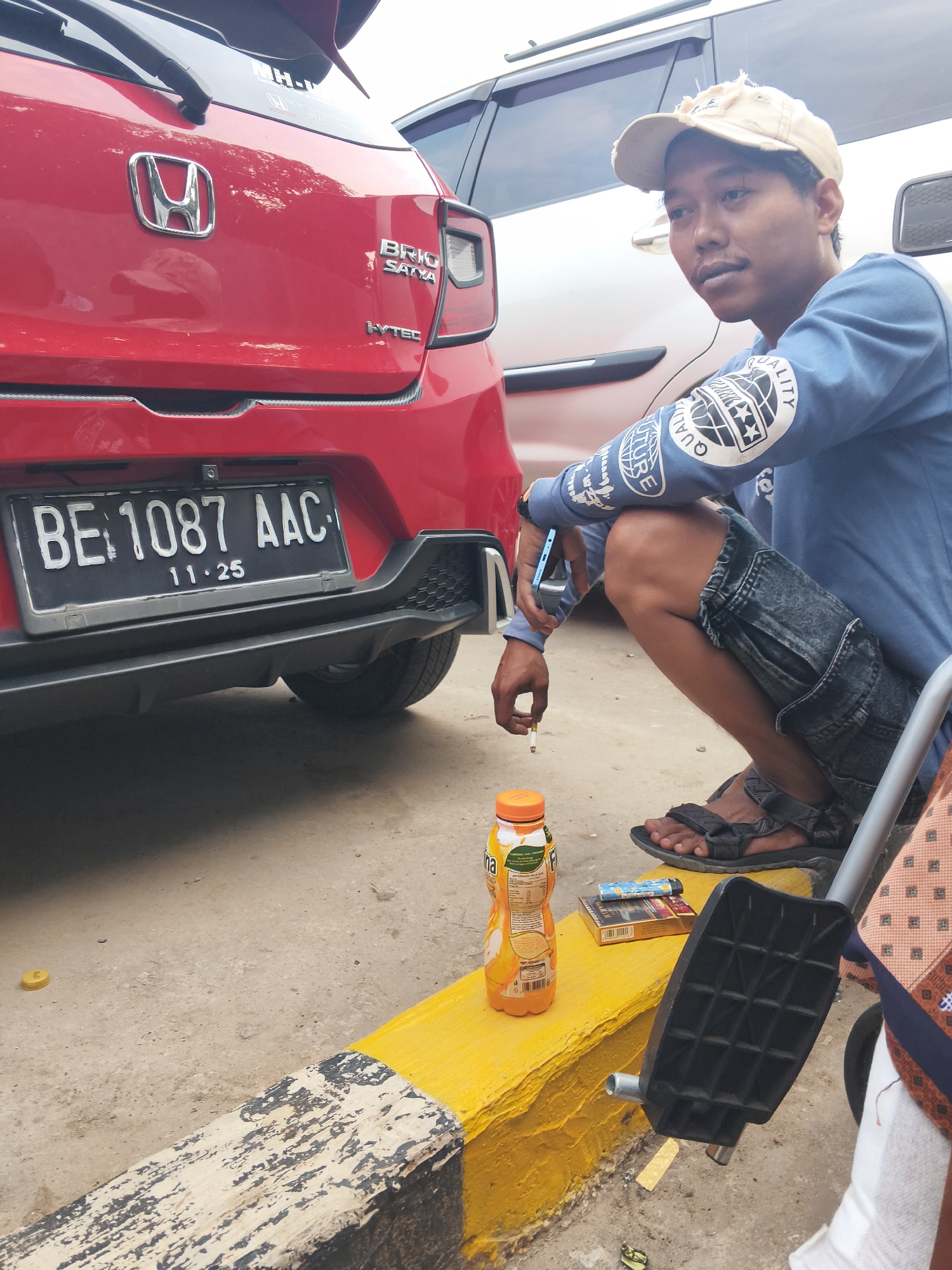 A young man, appearing to be of Indonesian descent, is squatting beside a red car with a license plate reading "BE 1087 AAC". He is wearing a blue long-sleeved shirt with graphics on the sleeves, denim shorts, a beige baseball cap, and dark sandals. In his right hand, he holds a lit cigarette, and his left hand rests on his knee.
In front of him, on a yellow and black striped curb, are a bottle of orange juice and a pack of cigarettes with a lighter. The car is a Honda Brio Satya, indicated by the badging on the trunk. The background shows another car and a slightly hazy sky. The scene appears to be set on a street or parking lot in Bandar Lampung, Indonesia, given the license plate format which is typical for Lampung province. The ground is paved with asphalt and some dirt.