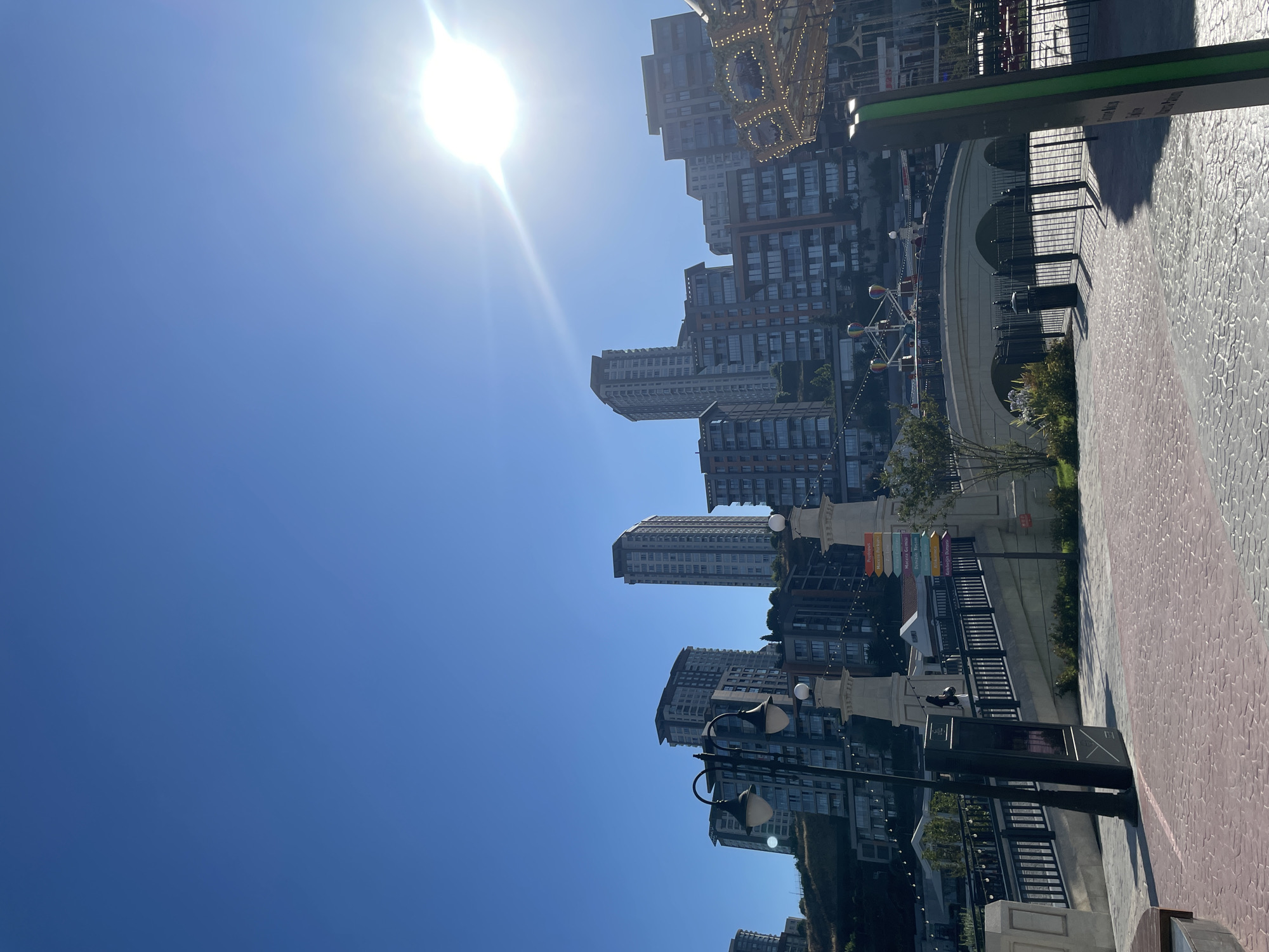 This is an outdoor, eye-level shot of a sunny day in Küçükçekmece, Turkey, with a clear blue sky and bright sun, partially obscured by a lens flare. The scene features a modern urban landscape with several multi-story apartment buildings visible on a hillside. In the foreground, a paved walkway with a red and white patterned brick surface leads towards a bridge. The bridge has decorative white pillars and black metal railings, with a ramp ascending from the left side. A lamppost with two globes stands to the left of the walkway. Behind the bridge, a small amusement ride with colorful balls and lights is in operation, and further back, a carousel structure is partially visible. A directional signpost with multiple colored signs in Turkish is located to the right of the frame. A few individuals are seen walking on the bridge, and trees and bushes are scattered throughout the scene. The overall atmosphere is bright and lively, suggesting a public recreational area.