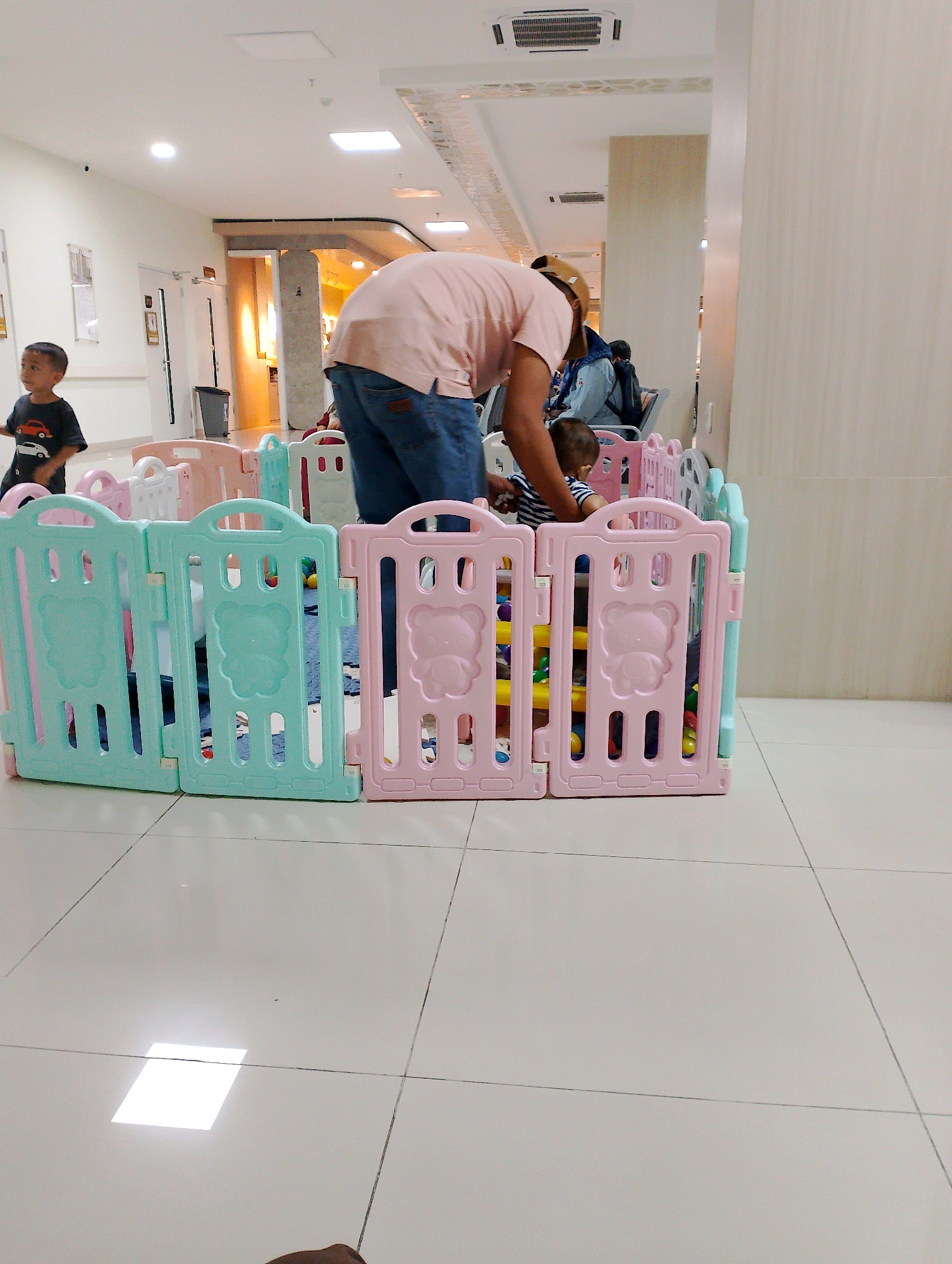 A pediatric ward waiting area in Kebonagung, Indonesia, features a playpen constructed from pastel pink and mint green plastic panels, containing colorful balls. A man in a light pink polo shirt and blue jeans is bent over, attending to a baby inside the playpen. To the left of the playpen, a young boy in a black t-shirt with a car print stands looking to the left. The room has a tiled floor and a drop ceiling with recessed lighting. In the background, a person sits on a chair.