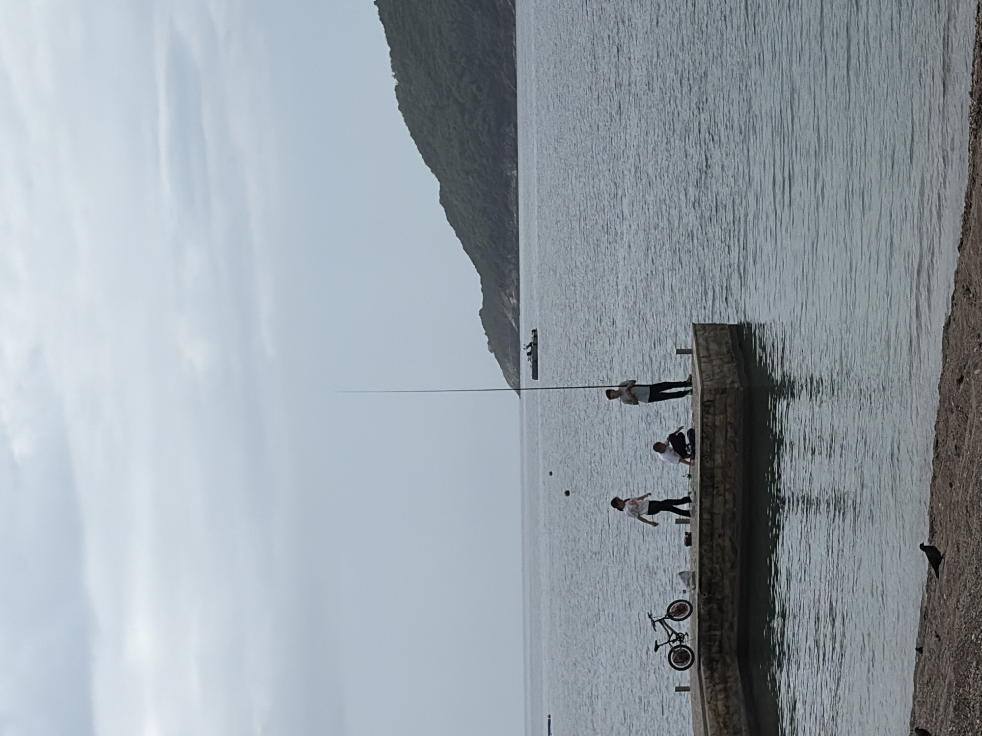 The image captures a waterfront scene in Budva, Montenegro. A gray, overcast sky provides diffuse lighting. A large mountain dominates the left side of the scene. A calm body of water stretches across the majority of the image. On a concrete pier that extends into the water, three people are present. One person stands near the end of the pier holding a fishing rod with a very long line. Another person sits on the pier further down. A third person walks near a bicycle that's leaning against the pier. The pier abuts a beach with dark sand and a scattering of rocks.