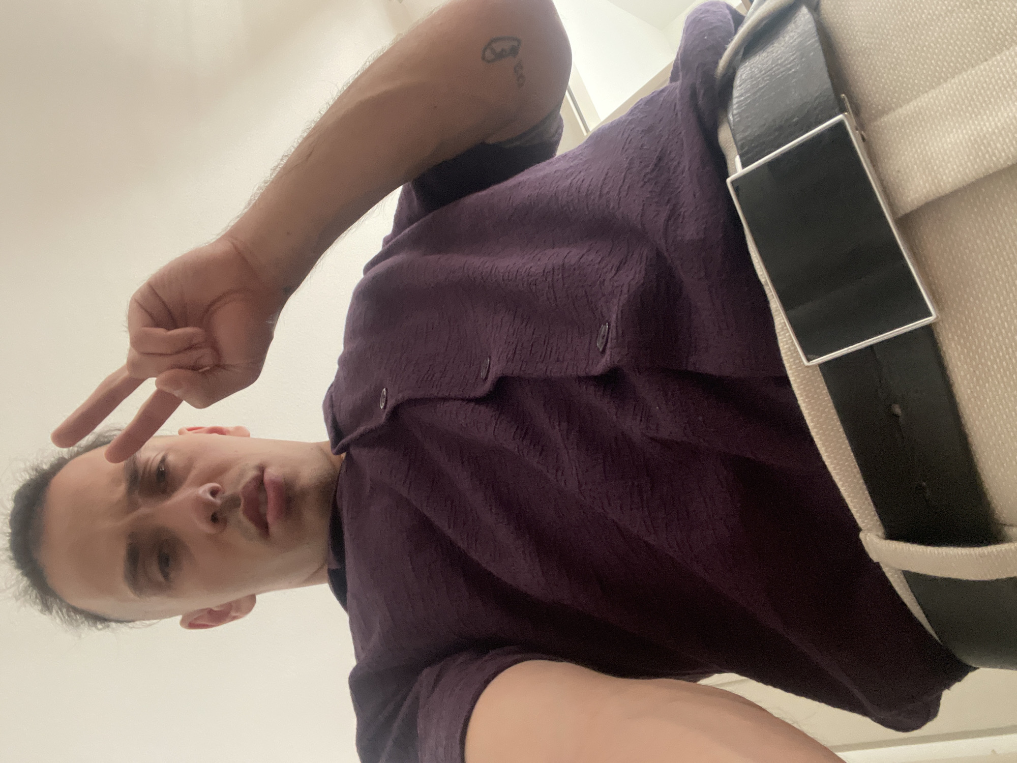 A person is taking a selfie indoors with a white wall as the background. The person is wearing a dark purple button-down shirt and a black belt with a rectangular buckle over beige pants. The person is holding up two fingers in a peace sign. The location is Unknown, Unknown.