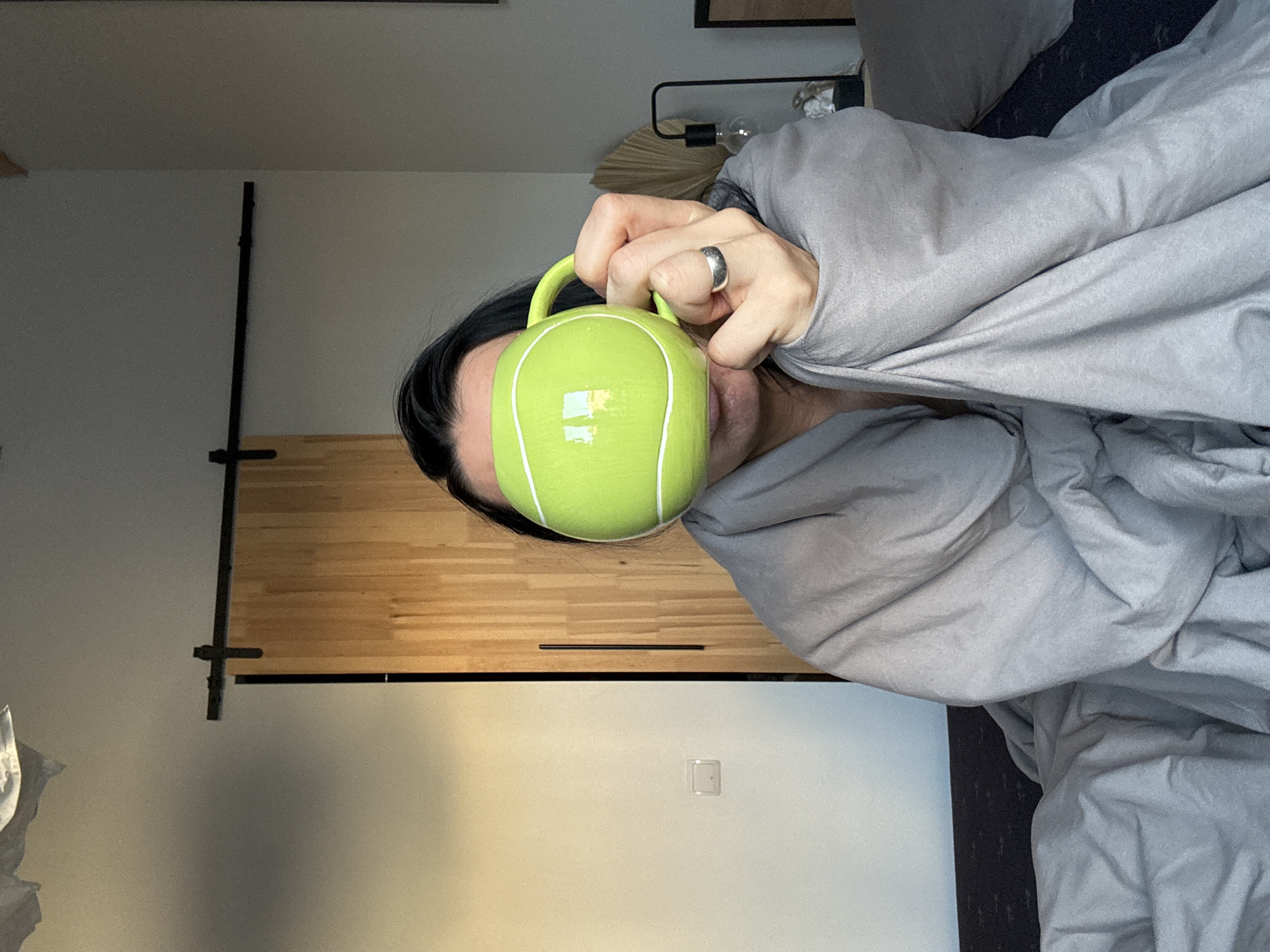 A person lies in bed under a gray comforter and holds a bright green tennis ball-shaped mug in front of their face. The person is wearing a ring on their right hand. The background shows a bedroom with a sliding wooden door. A light fixture is visible on the wall behind the bed. The location is most likely in a home, and while a city is provided, the specific location within Chaikovsky, Russia, cannot be determined from the image.