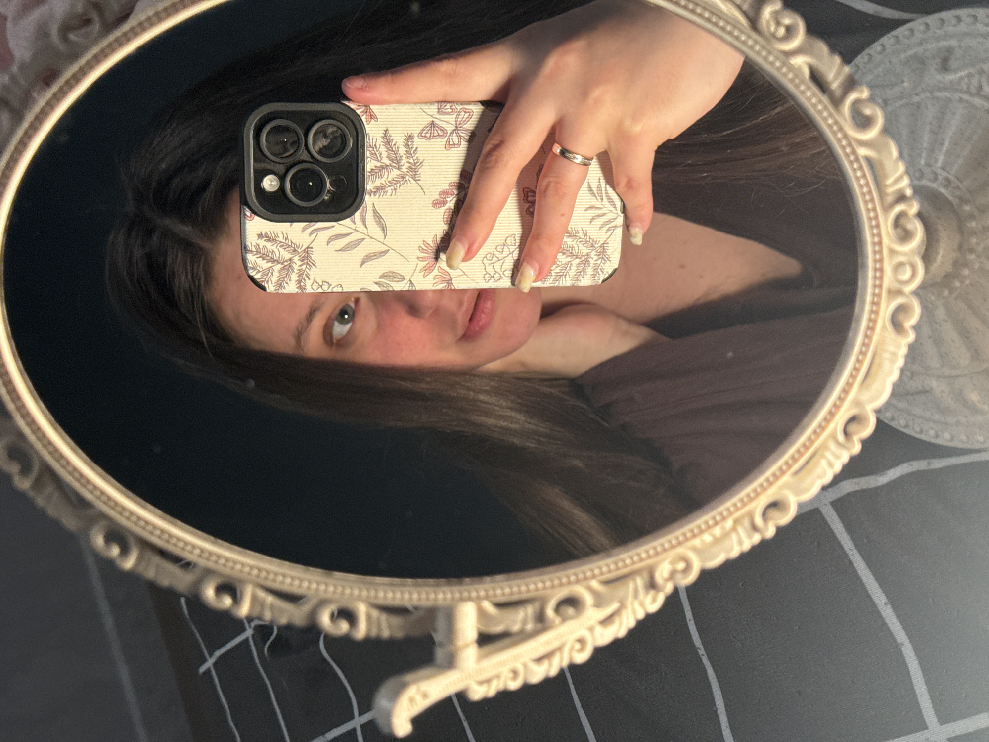 A person is reflected in an oval mirror with an ornate off-white frame. The person is holding a smartphone up to the mirror, obscuring the upper half of their face. The phone has a triple-lens camera and a case with a floral and butterfly pattern. The person wears a ring on their finger. The mirror is on a stand, and below the mirror is a dark, plaid surface. To the right of the mirror, out of focus, is a white object. There is no information to determine the location is in Krasnodar, Russia.