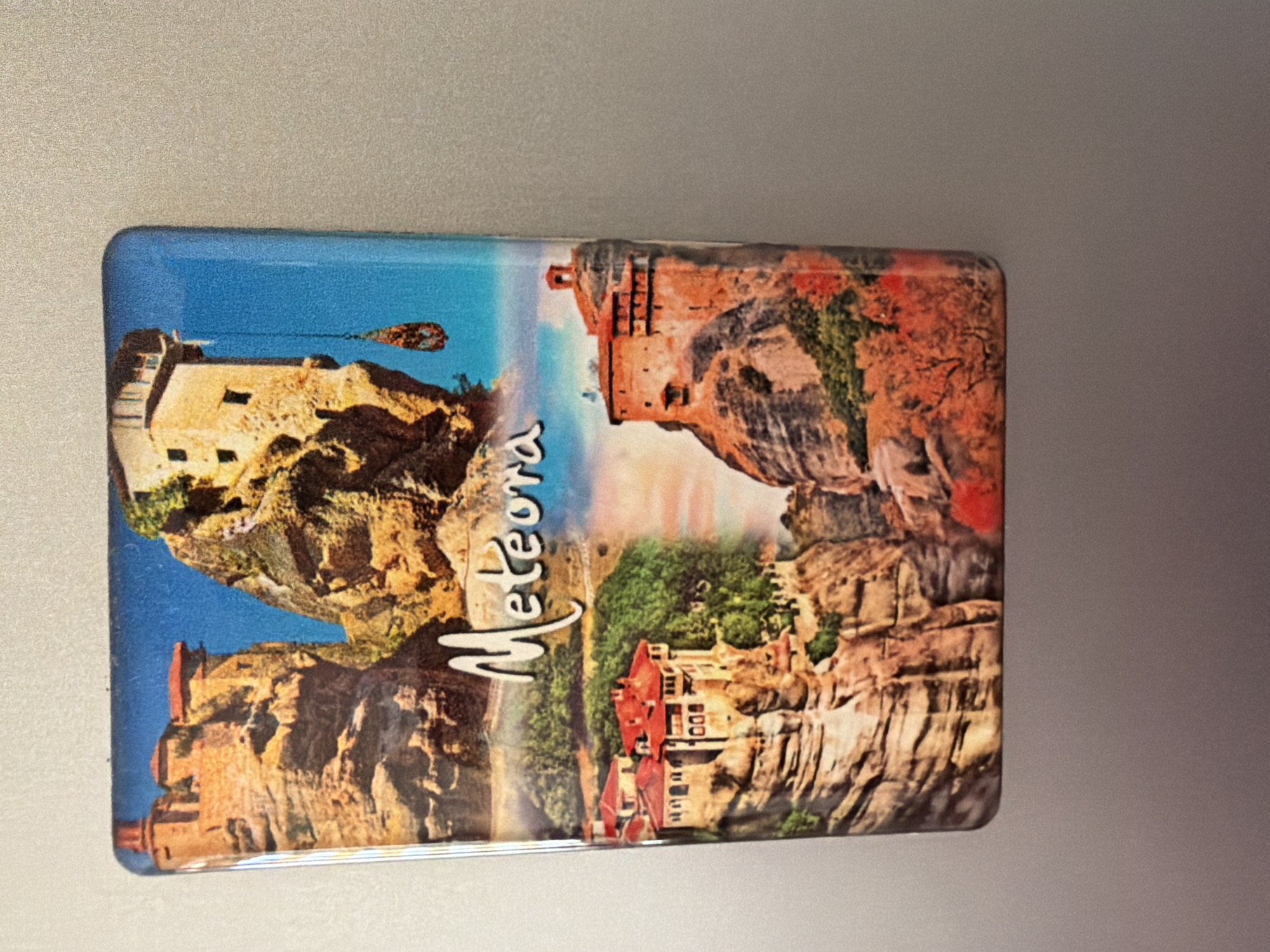 The image is a close-up, slightly angled shot of a rectangular magnet with a colorful depiction of the Meteora monasteries in Greece. The magnet is placed on a light gray, textured surface, likely a refrigerator. The magnet is divided into sections, showing different views of the monasteries perched on towering rock formations. The top section features a monastery with red roofs and stone walls, set against a bright blue sky. Below this, the word "Meteora" is written in a flowing white script across a band of colorful clouds. The bottom section of the magnet displays two more views of monasteries. One shows a complex of buildings with red tiled roofs nestled on a rocky outcrop amidst lush green trees. The other, to the right, shows a monastery with an orange-hued facade built directly onto a steep, rocky cliff face, with autumnal trees in shades of red and orange at its base. The magnet's surface is glossy, and there are subtle reflections on it. There is no presence of the city of Pazardzhik, Bulgaria in this image.