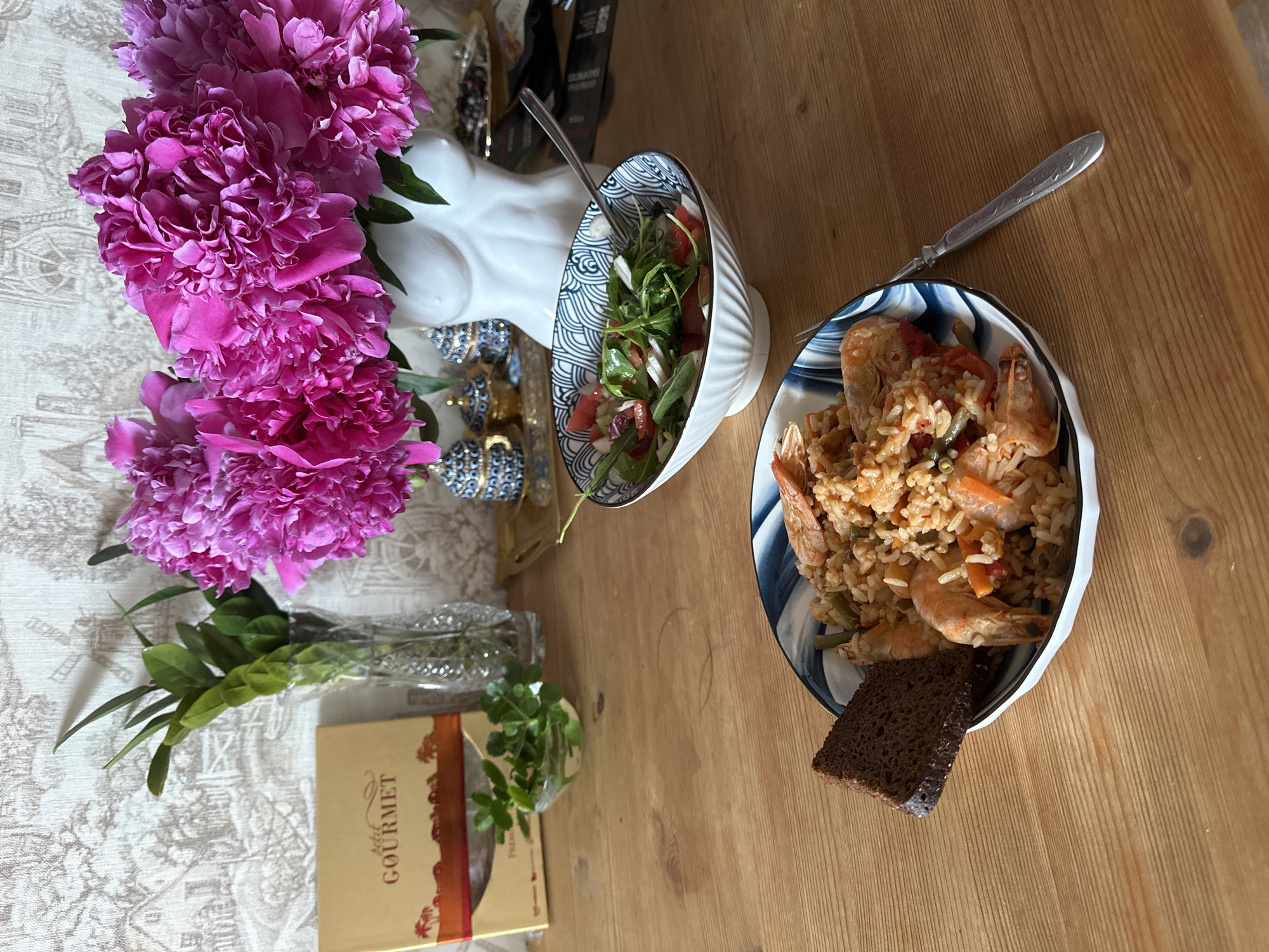 The image depicts a wooden table set with a meal. In the foreground, a bowl of rice and shrimp is garnished with a slice of dark bread. To the right, a bowl of salad with greens and tomatoes is placed, with a fork resting on the edge. In the background, a bouquet of vibrant pink peonies is arranged in a white bust-shaped vase. A glass vase holding green foliage stands to the left of the peonies, alongside a golden box labeled "GOURMET." A decorative wallpaper with a pastoral scene serves as the backdrop. The overall setting suggests a meal enjoyed indoors. There is no indication of the city of Vladimir, Russia, within the image.