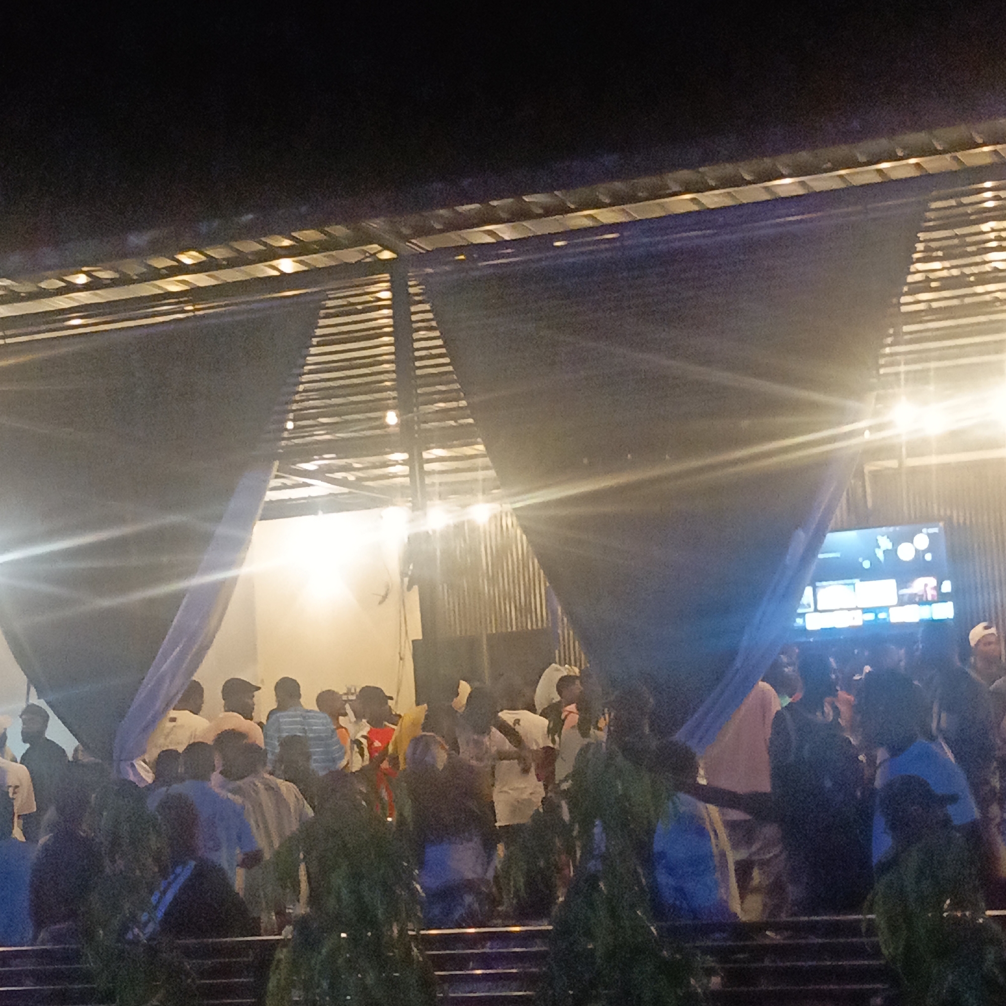 A crowd of people is gathered under a corrugated metal roof structure with decorative black fabric and blue accents, likely at night given the low light and visible artificial lights. The location is presumably Ube, Nigeria. Many individuals are wearing casual clothing, including hats and caps, and appear to be standing on steps. A screen displaying images is visible in the background. Tall plants are situated at the foreground near the steps.