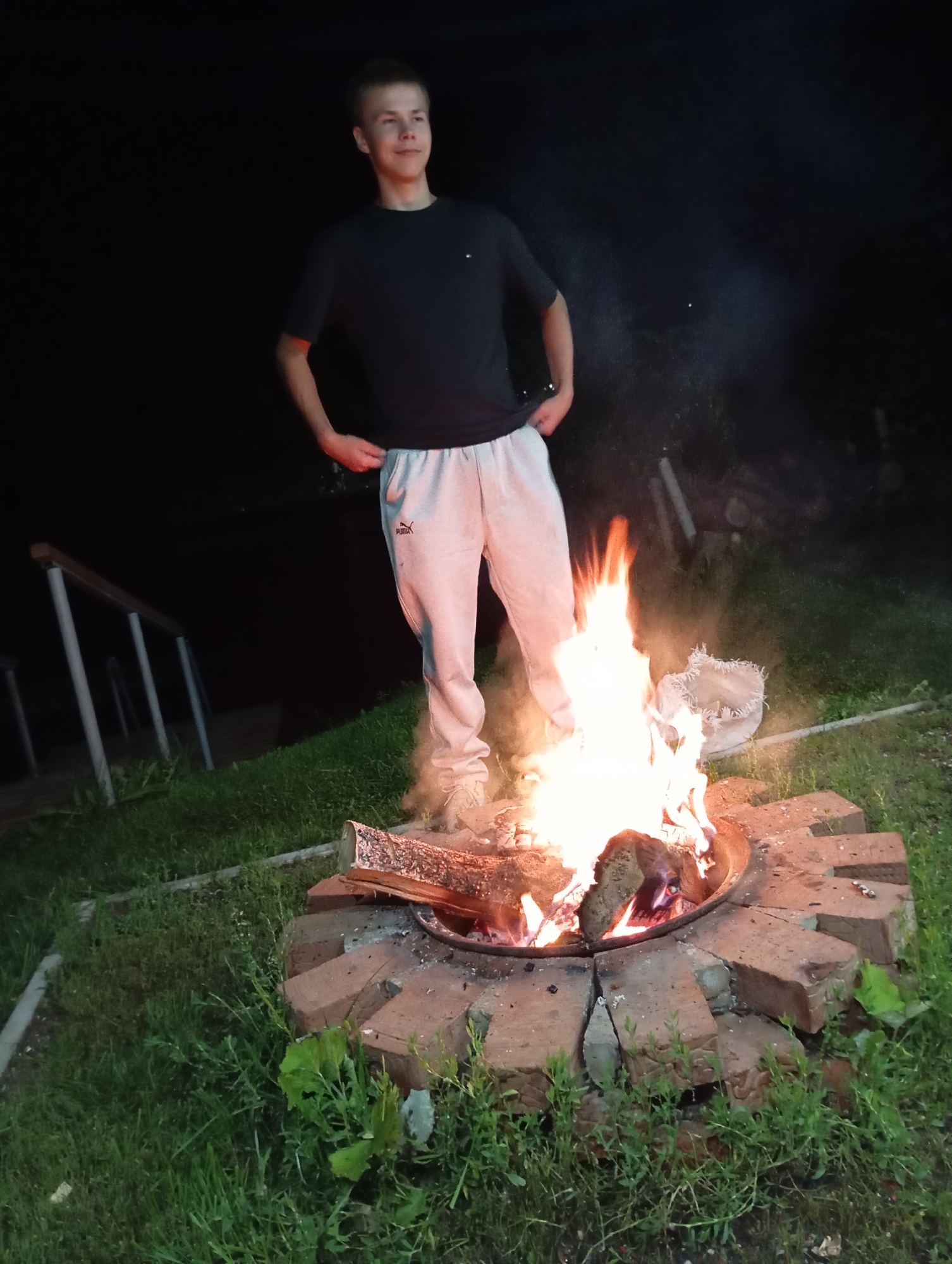 A young male stands near a campfire at night. He is wearing a black t-shirt and light grey sweatpants. The campfire is made of wood and is contained in a brick fire pit. The background is dark and appears to be an outdoor setting, possibly a backyard or park, with grass visible around the fire pit. There are some wooden steps or railings to the left of the frame. The overall impression is of a casual evening gathering around a fire. The location is identified as Malyi Bukor, Russia.