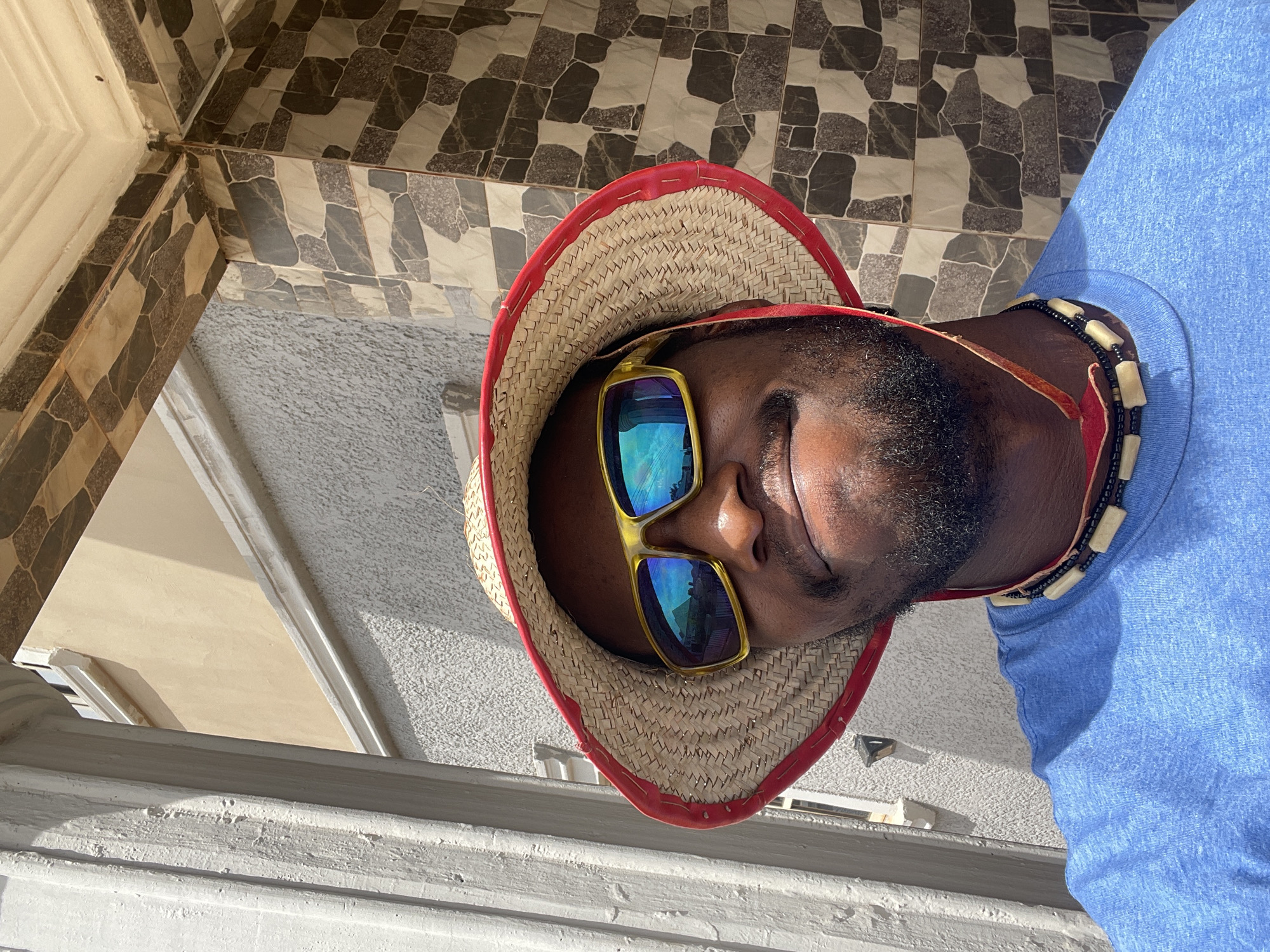 The original image is rotated 90 degrees clockwise, depicting a close-up, low-angle portrait of a man, likely taken looking upwards. The man has dark skin and a short beard, with his face partially obscured by bright yellow-framed sunglasses featuring reflective blue lenses. He is wearing a straw hat with a distinctive red fabric trim and a light blue t-shirt. Around his neck, a necklace made of small black beads interspersed with larger, light-colored rectangular beads is visible.
The background features an architectural setting, with a ceiling or upper wall covered in patterned tiles. These tiles display irregular geometric shapes in shades of dark grey, light grey, and beige, creating a mosaic-like effect. Other visible structural elements include sections of cream-colored and textured white or light grey walls, along with architectural moldings or ledges. The scene is brightly lit by natural sunlight, casting noticeable shadows, which indicates a daytime setting. The environment suggests an indoor or sheltered outdoor space, such as a porch or corridor, consistent with architecture found in Abuja, Nigeria. There is no visible text in the image.