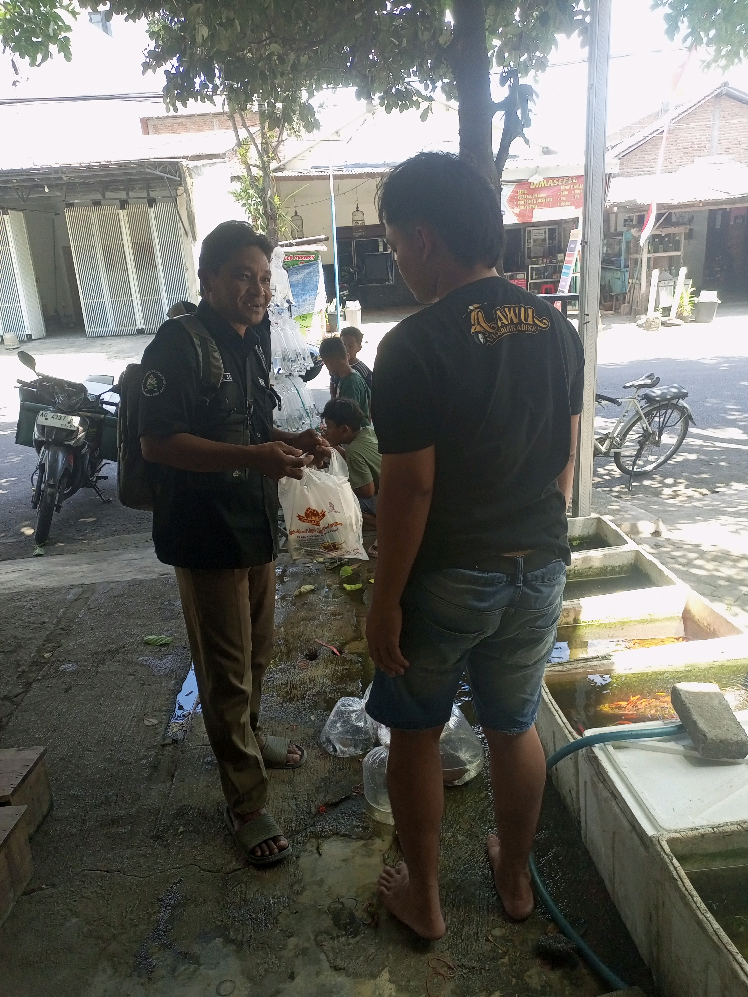 The image depicts an outdoor scene, likely in Kediri City, Indonesia, with two men interacting. The man on the left, wearing a dark shirt and khaki pants, holds a white plastic bag and appears to be speaking to the other man. He has a backpack on. The man on the right, wearing a black t-shirt with a graphic and denim shorts, faces away from the camera, showing the back of his t-shirt. He is barefoot. In the background, there are several tanks containing fish, suggesting the location might be a fish farm or market. A motorcycle is parked to the left of the frame. The environment is wet and appears to be paved with concrete, with trees providing shade overhead. Buildings and structures are visible in the distance. Several other individuals, including children, are partially visible in the background.