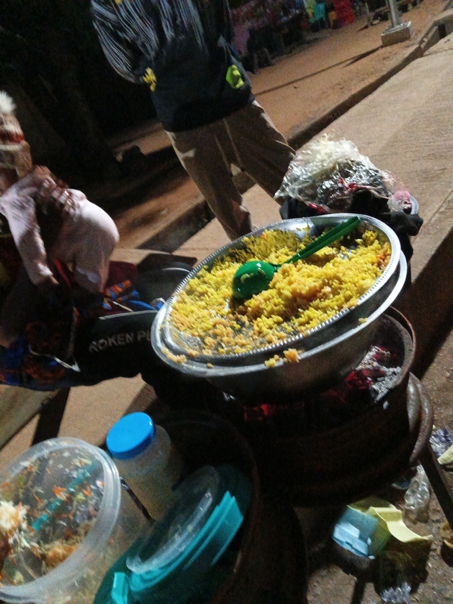 In Bukuru, Nigeria, an outdoor street food vending scene unfolds at night. A person, partially visible and wearing a dark shirt with a yellow emblem and brown trousers, stands near a large metal bowl filled with yellow rice. A green plastic ladle is submerged in the rice. The bowl rests on a makeshift stove, with glowing embers visible beneath it. In the foreground, several containers are present: a clear plastic container with food, a bottle with a blue cap, and another teal container. To the right, a plastic-wrapped bundle of food is placed on the ground. The ground appears to be paved, and the background shows other vendors or stalls with colorful items, illuminated by artificial light. The overall impression is a bustling night market environment.